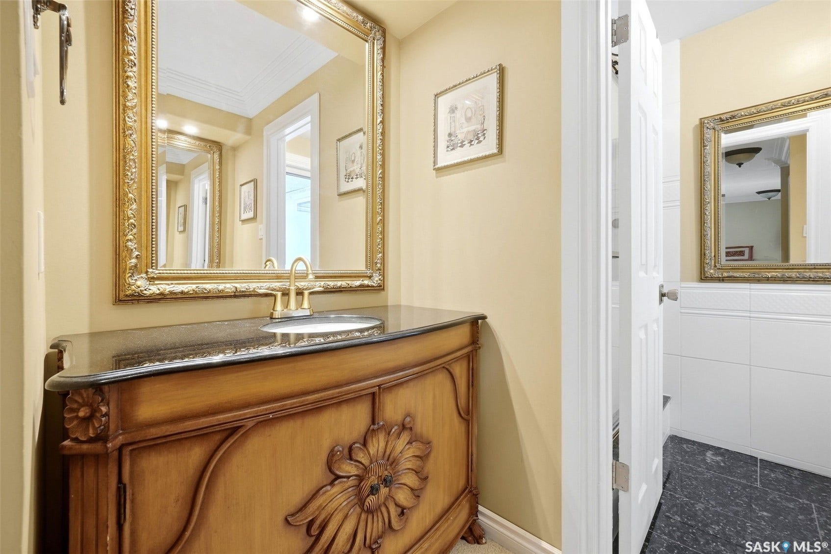 Ornate vanity with carved wood details, black countertop, gold-tone faucet, and a decorative framed mirror