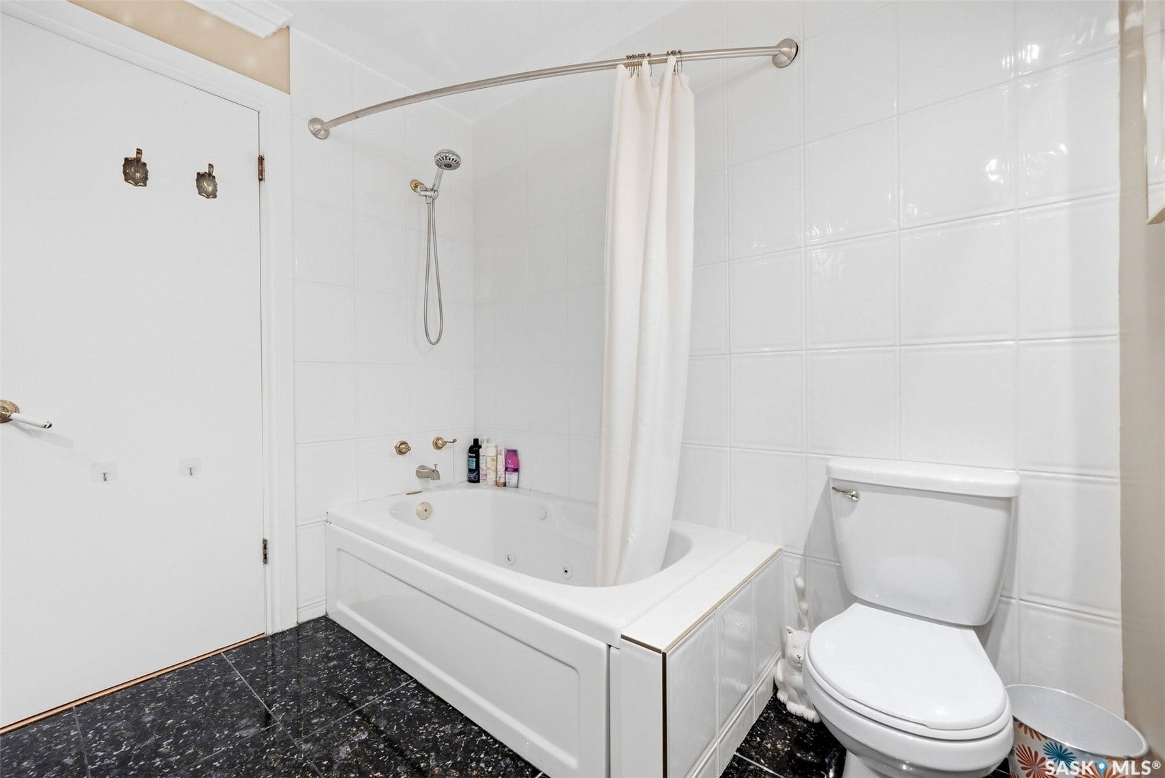 White tiled bathroom featuring a jetted tub, wall-mounted shower head, and dark speckled flooring