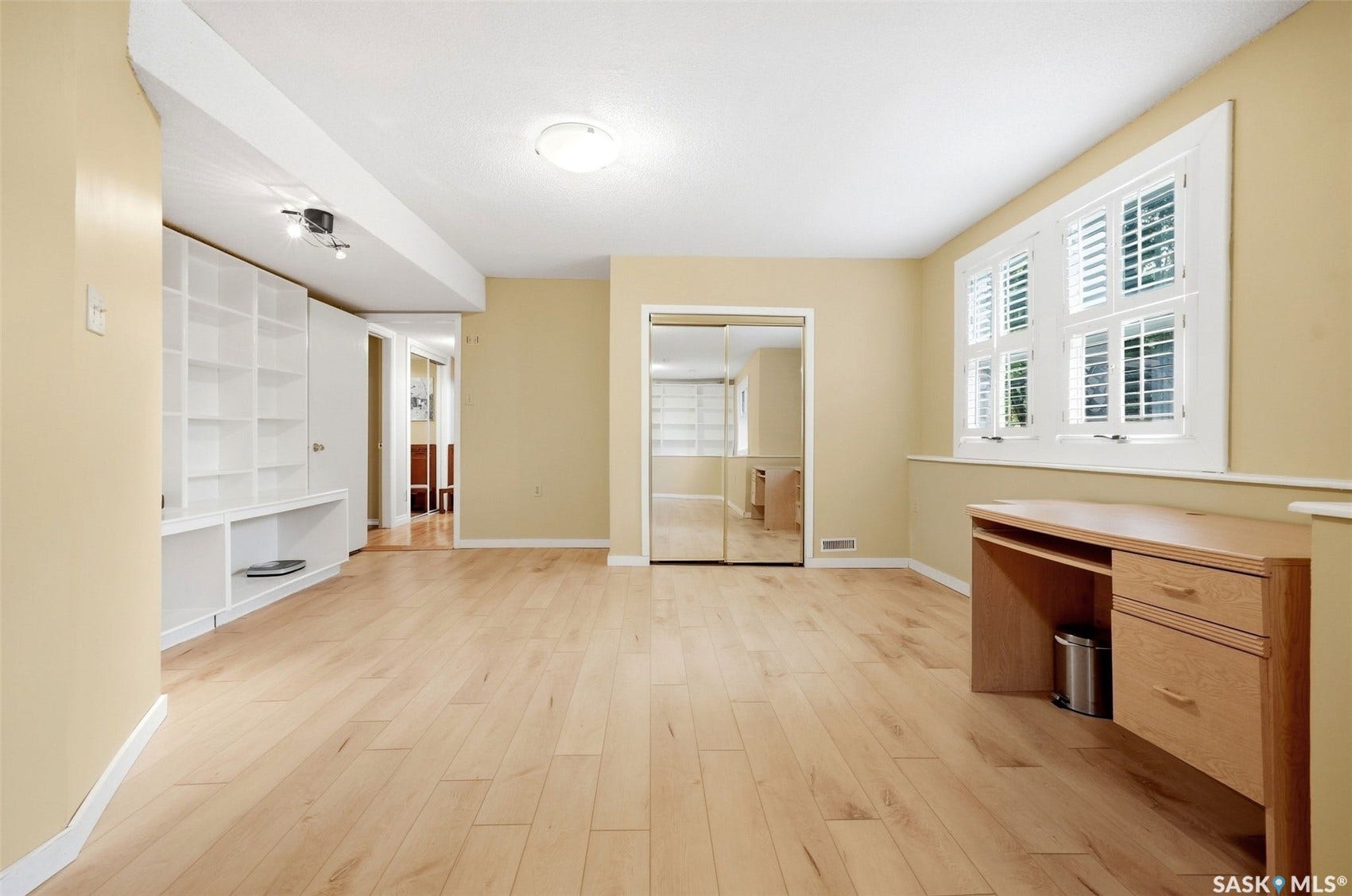 Light wood-finish flooring, built-in shelving, mirrored closet doors, and a window with white shutters