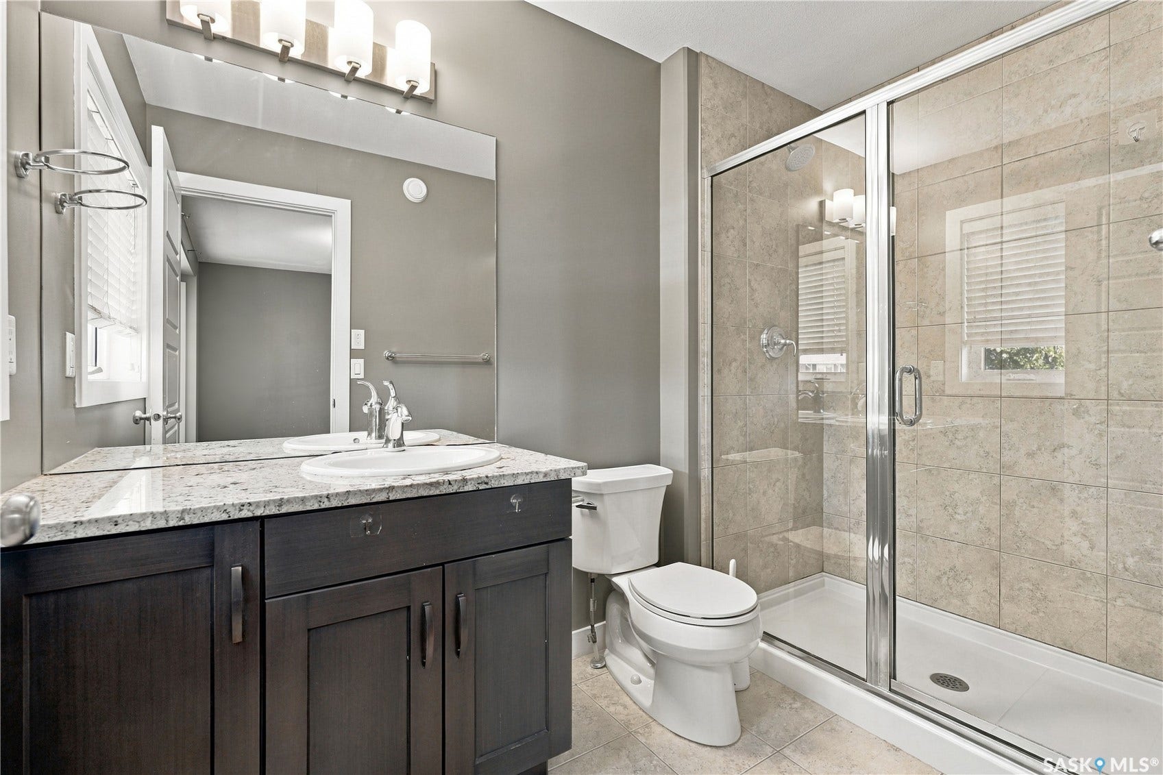 Bathroom featuring a dark wood-finish vanity, granite countertop, built-in sink with chrome faucet, a large frameless mirror, and a glass-enclosed shower with tiled walls