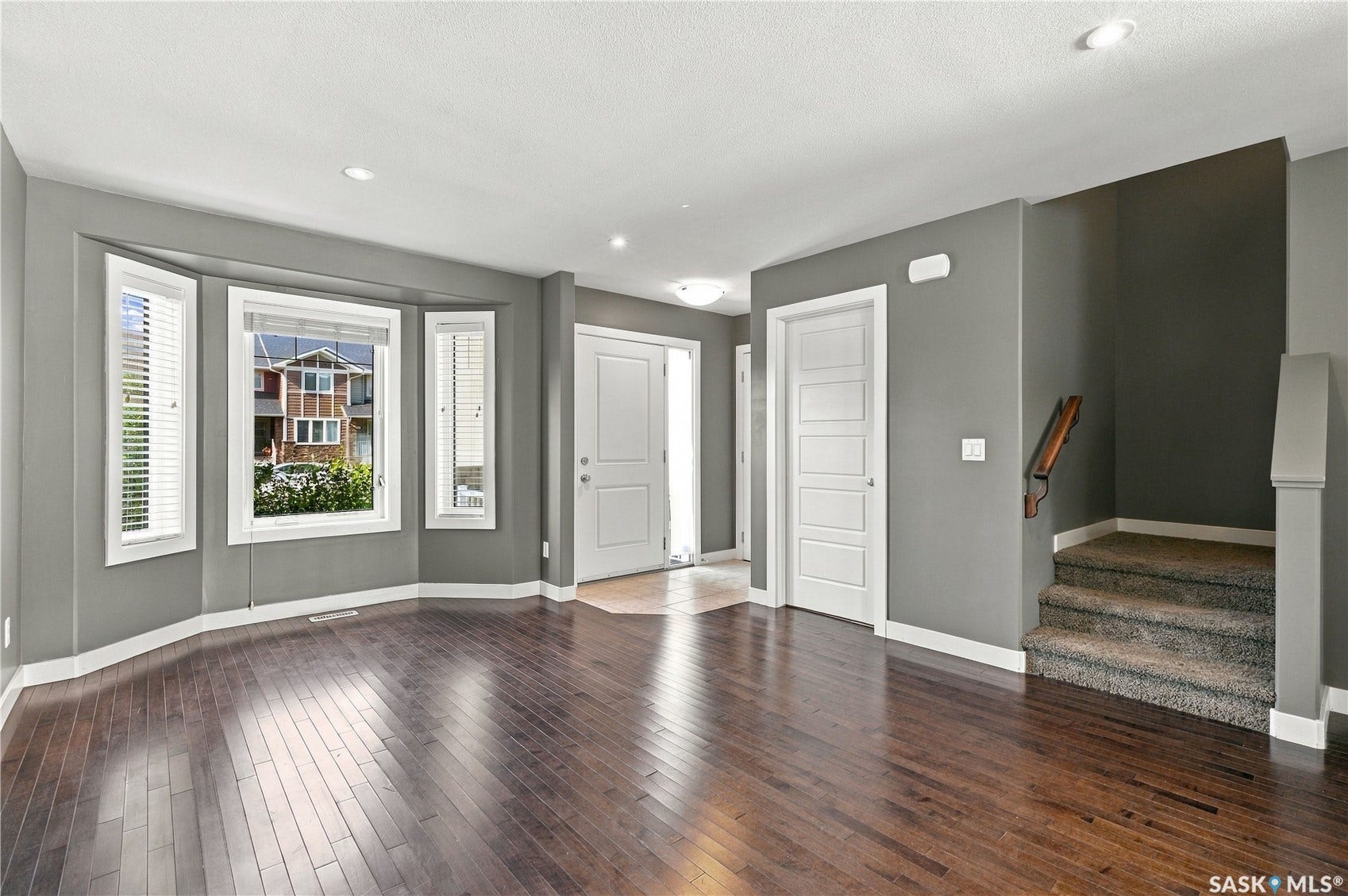 Bay window with white blinds, dark wood-finish flooring, recessed lighting, white paneled doors, and a carpeted staircase with a wood handrail