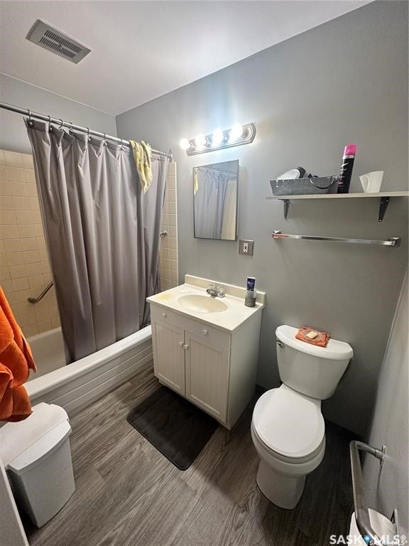 Bathroom featuring wood-finish flooring, a single vanity with storage, a toilet, a tub with an integrated shower, and wall-mounted shelving