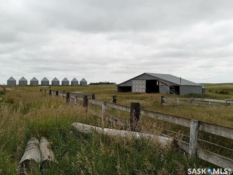 Expansive rural setting featuring a weathered wooden fence, a large metal barn structure, and a row of multiple grain silos