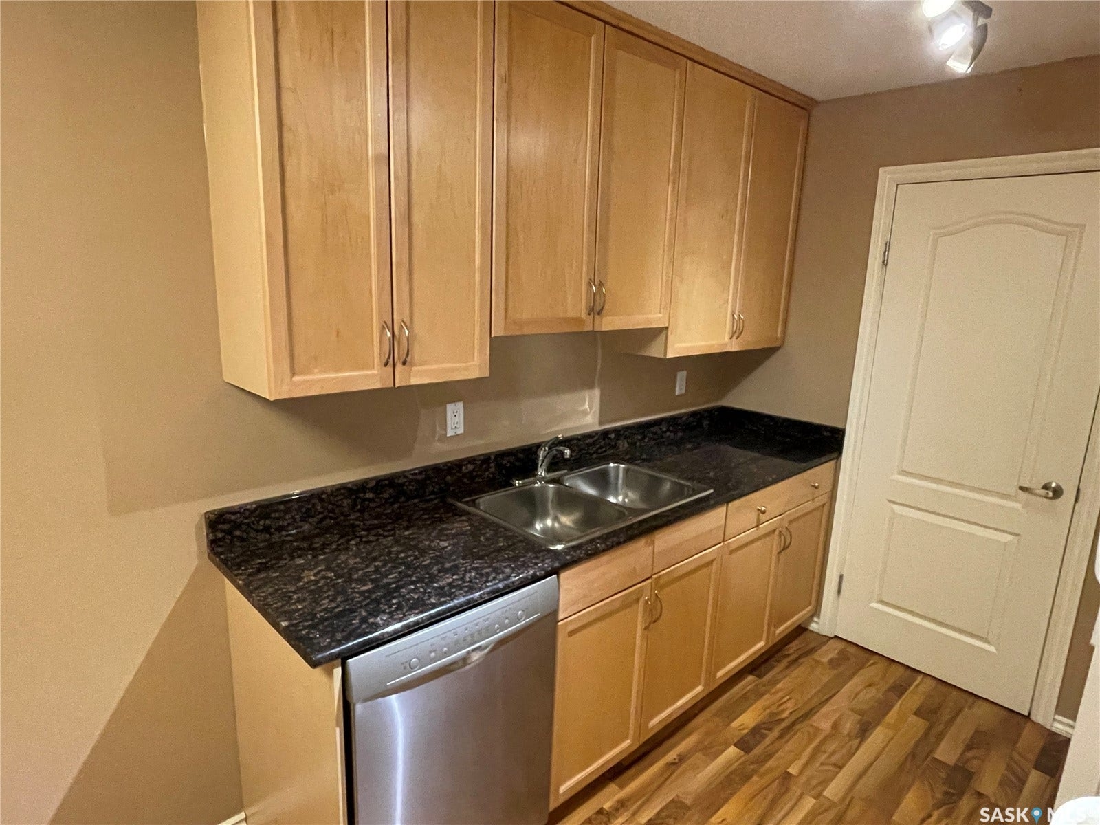 Light wood-finish cabinetry with silver-tone hardware, dark countertop with a double basin sink, stainless steel dishwasher, and wood-finish flooring