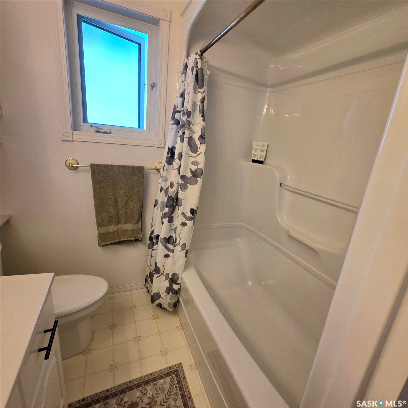 White acrylic tub and shower combination with an integrated grab bar, complemented by a white window frame and light-toned patterned floor tiles
