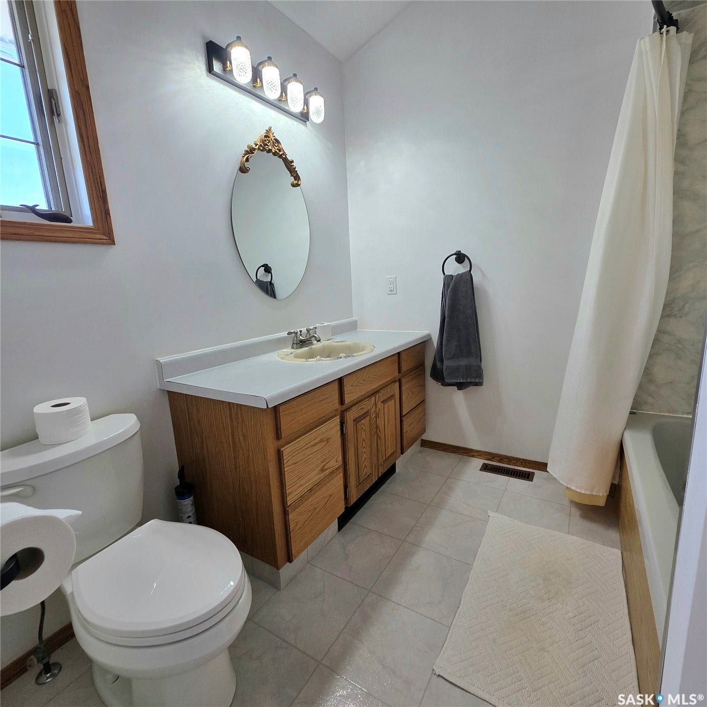 Vanity with wood-finish cabinetry and light-toned countertop, oval mirror with ornate frame, four-light fixture, white toilet, and tile flooring