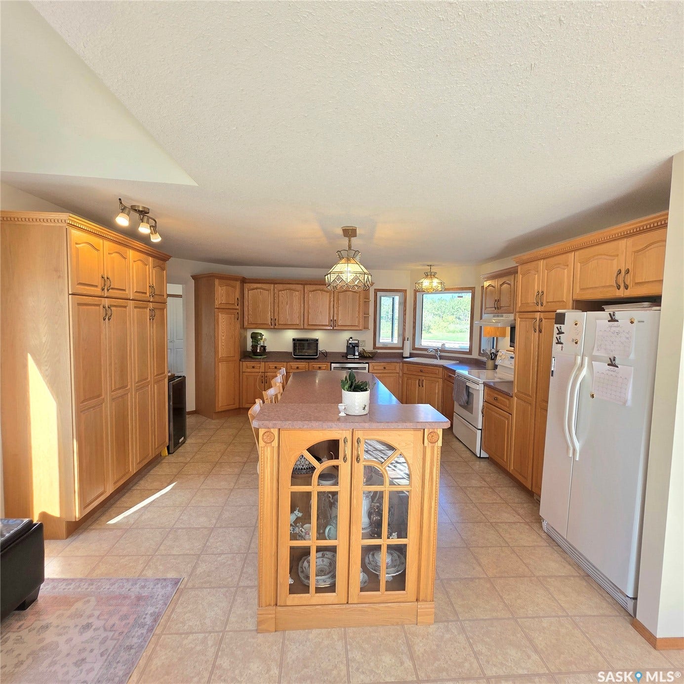 Spacious kitchen featuring extensive wood cabinetry, a large center island with built-in display cabinets, light-toned tile flooring, and multiple windows
