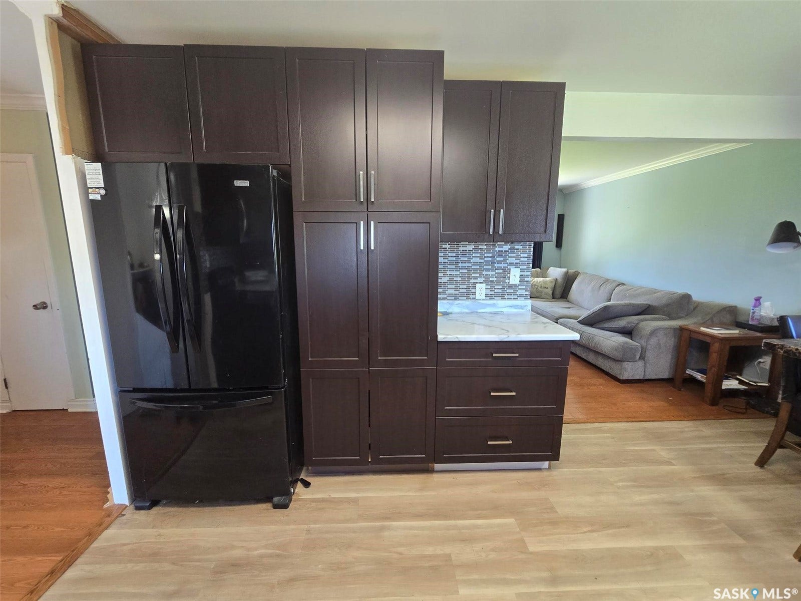 Dark wood-finish cabinetry with modern hardware, integrated black refrigerator, light-toned wood-finish flooring, white countertop, and mosaic tile backsplash