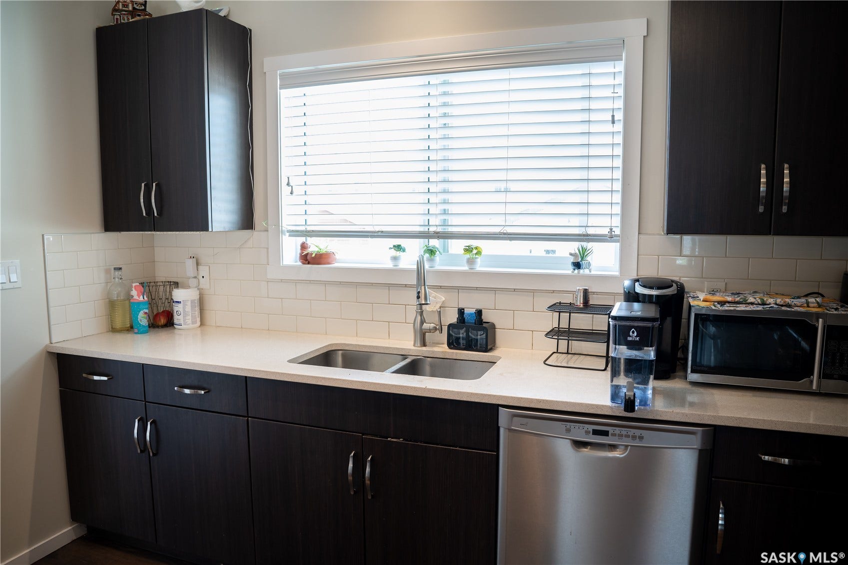 Dark wood-finish cabinetry, light-toned countertops, white subway tile backsplash, stainless steel double-basin sink, and a built-in dishwasher