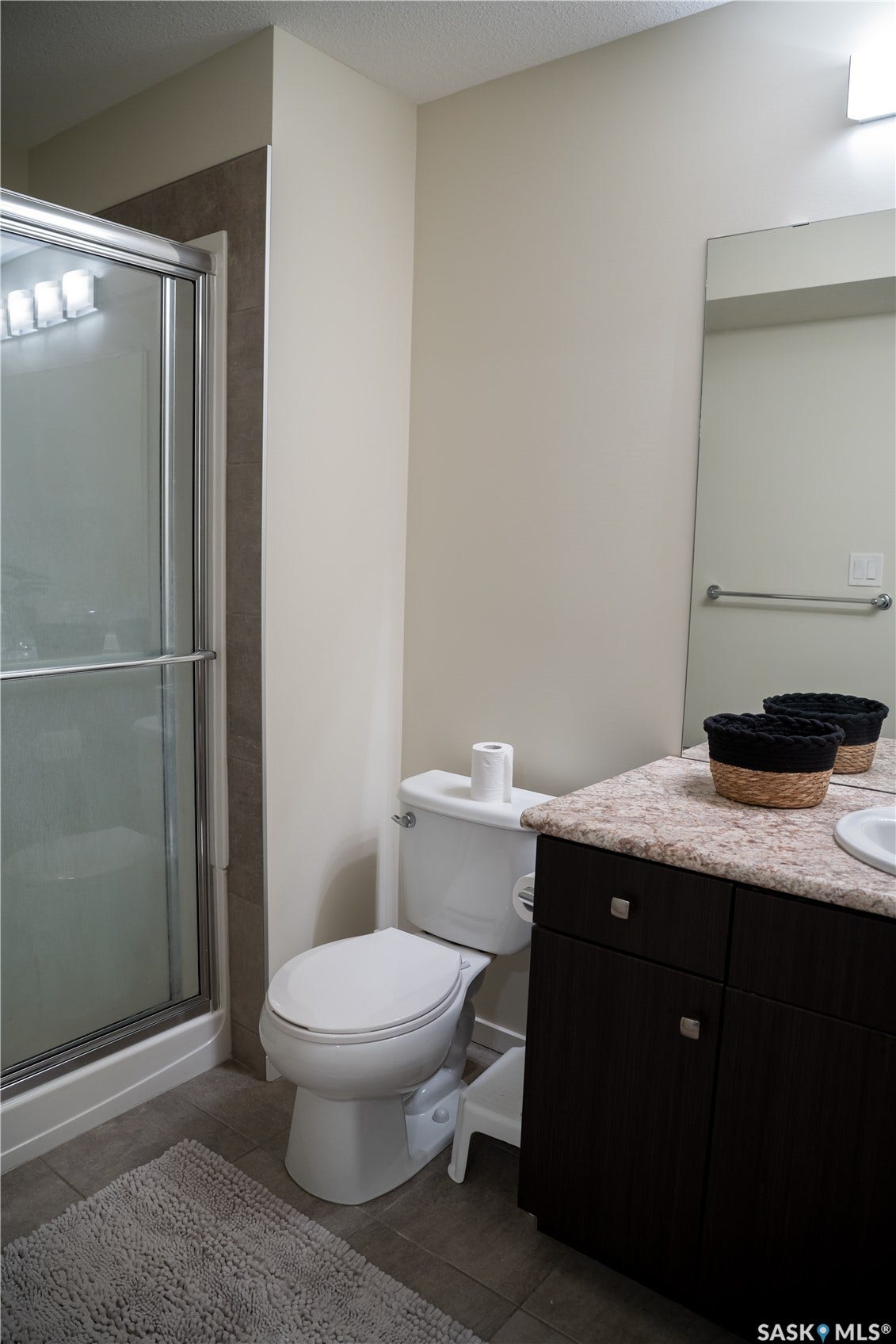 Shower with sliding glass enclosure, white toilet, dark wood-finish vanity with light-toned countertop, and a wall-mounted mirror