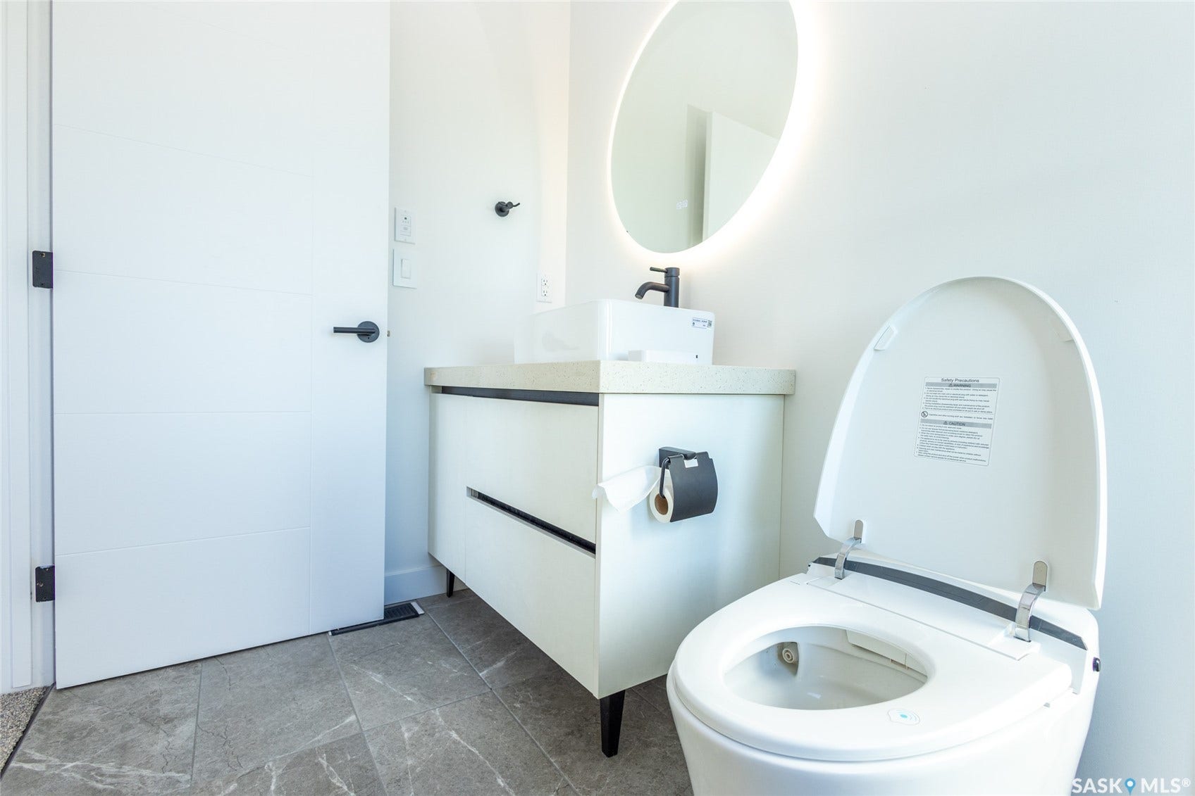 Modern bathroom featuring gray-toned tiled flooring, a white floating vanity with a light-colored countertop, and a circular back-lit mirror