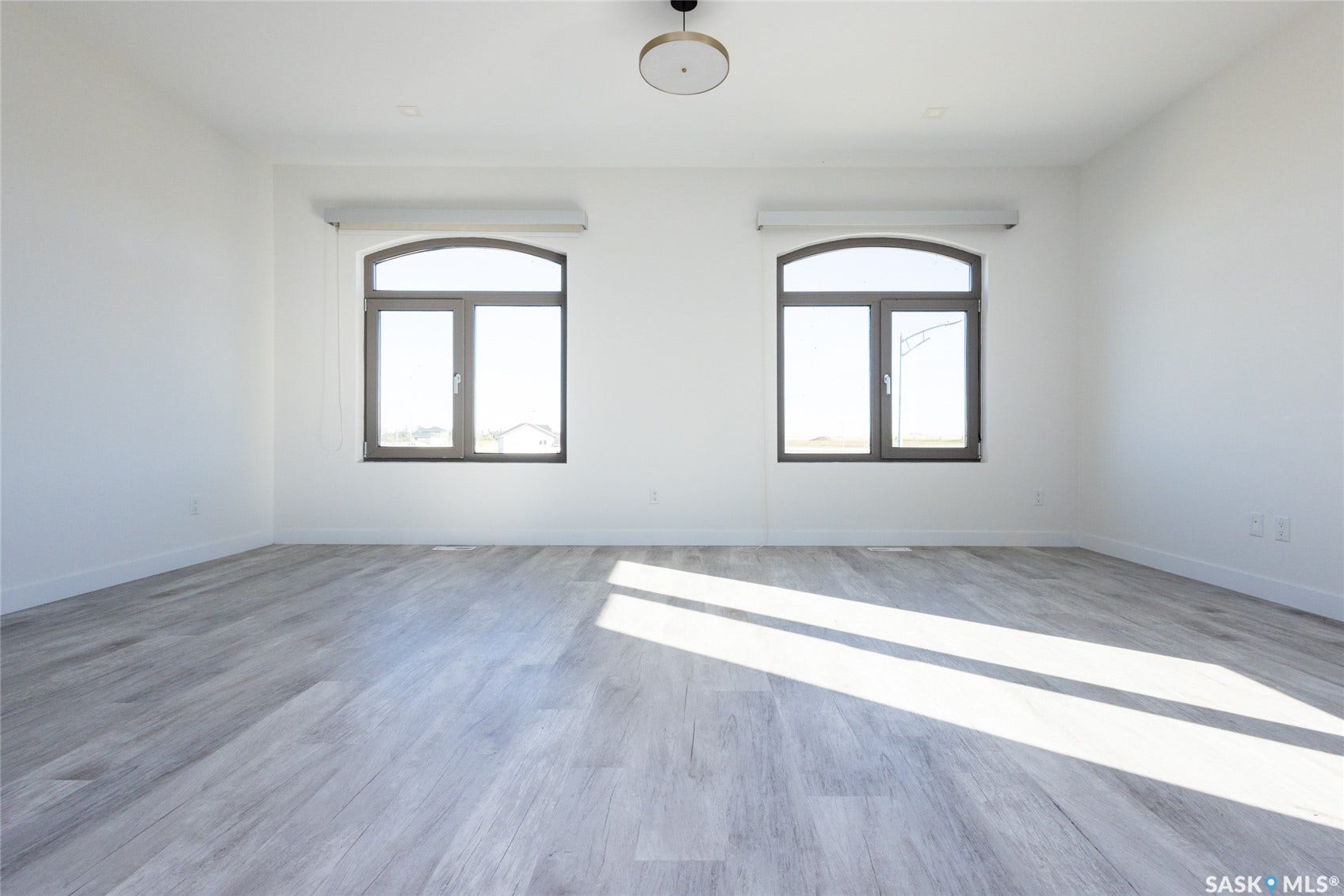 Spacious room featuring light wood-finish flooring, white walls, two arched transom windows, and a modern ceiling light fixture