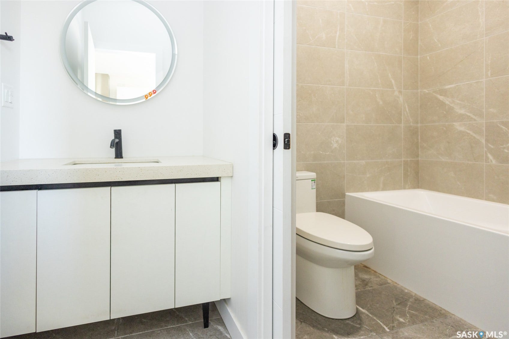 White vanity with integrated sink, matte black faucet, round mirror, and a built-in tub with tile surround