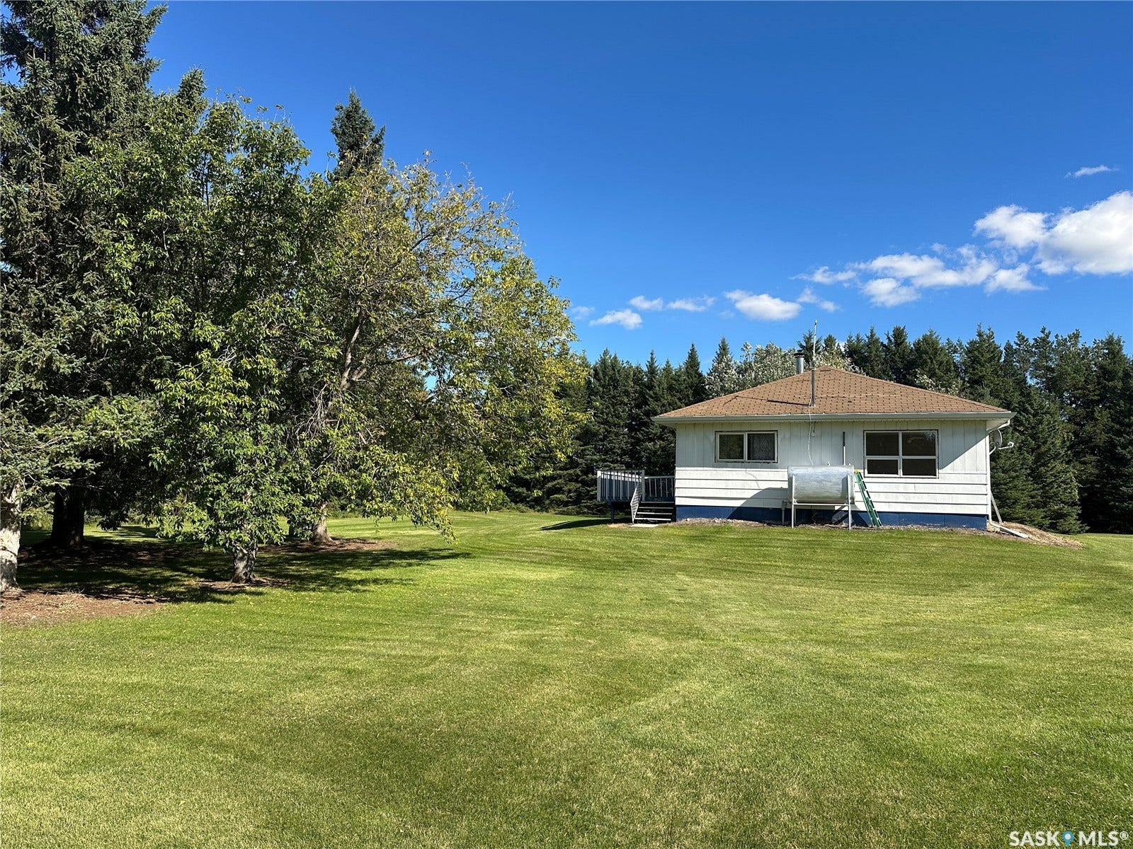 Expansive green lawn, mature trees, single-story residence with white siding, brown shingle roof, and a wood deck
