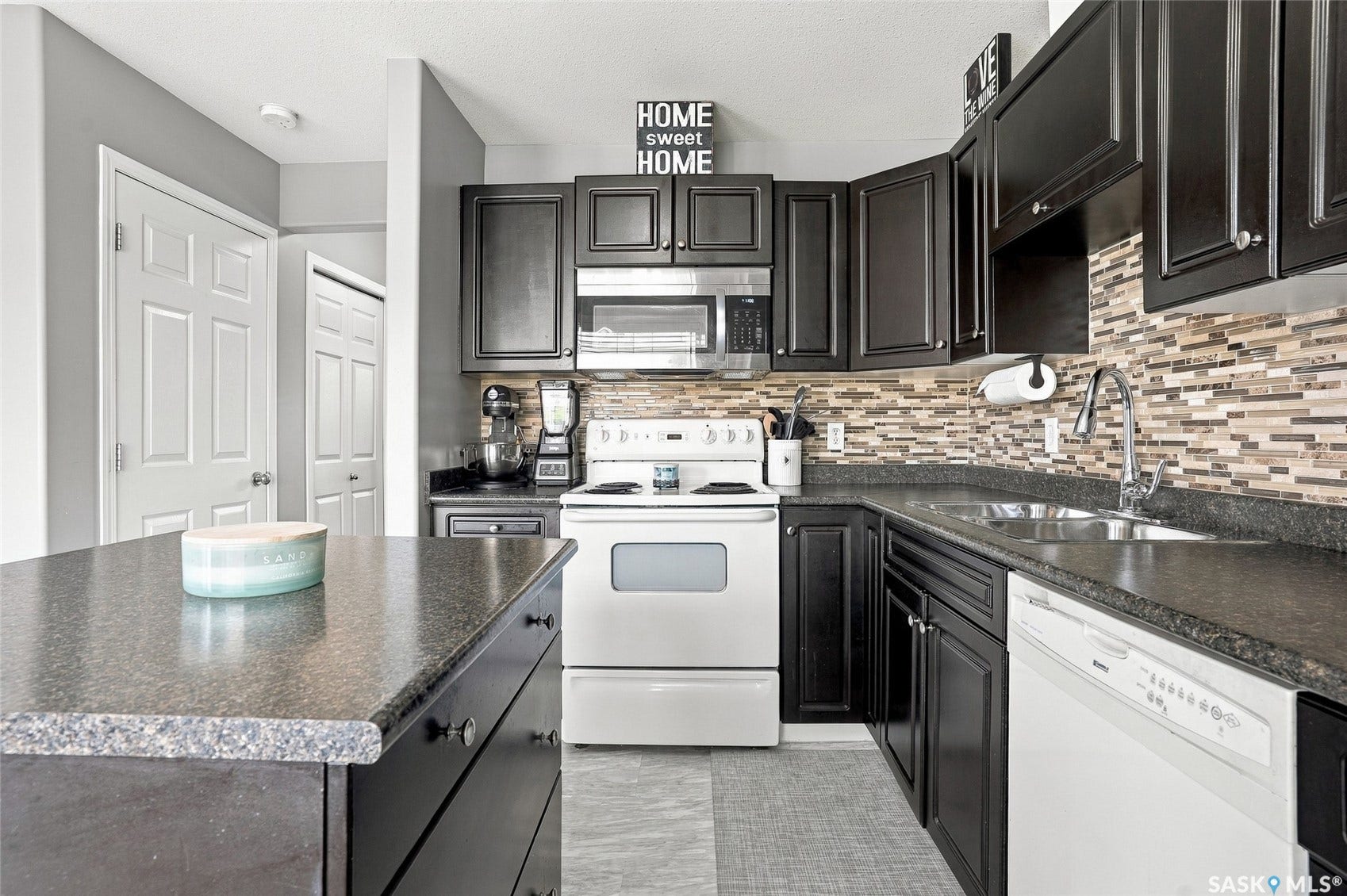 Dark cabinetry, white appliances, mosaic tile backsplash, double-basin sink, and a kitchen island with storage