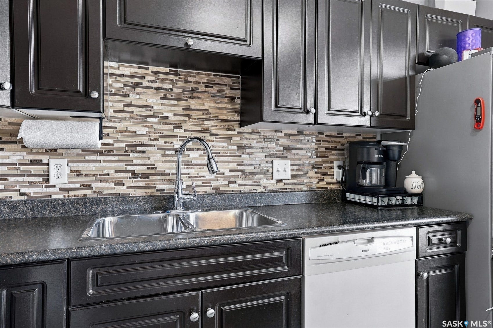 Dark wood-finish cabinetry, mosaic tile backsplash, stainless steel sink, chrome gooseneck faucet, and integrated dishwasher
