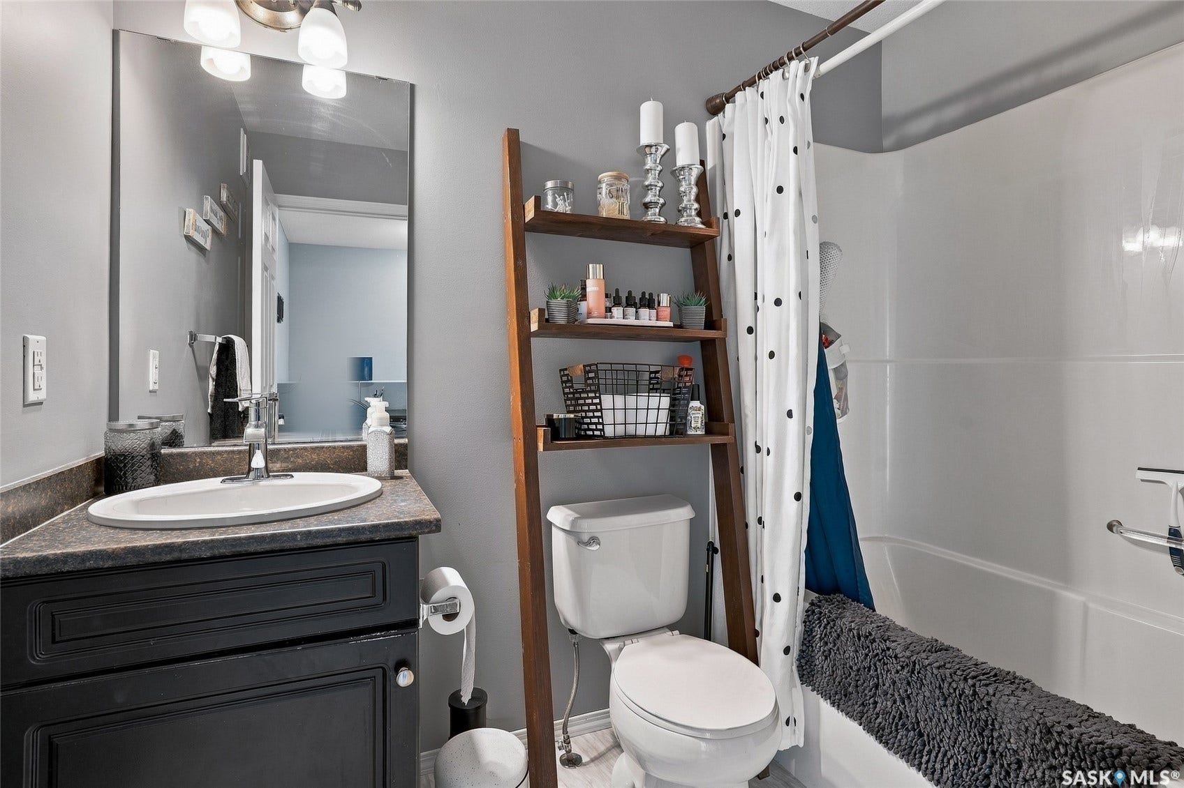 Vanity with dark cabinetry and a drop-in sink, framed mirror with overhead lighting, a white toilet, and a white tub with a shower