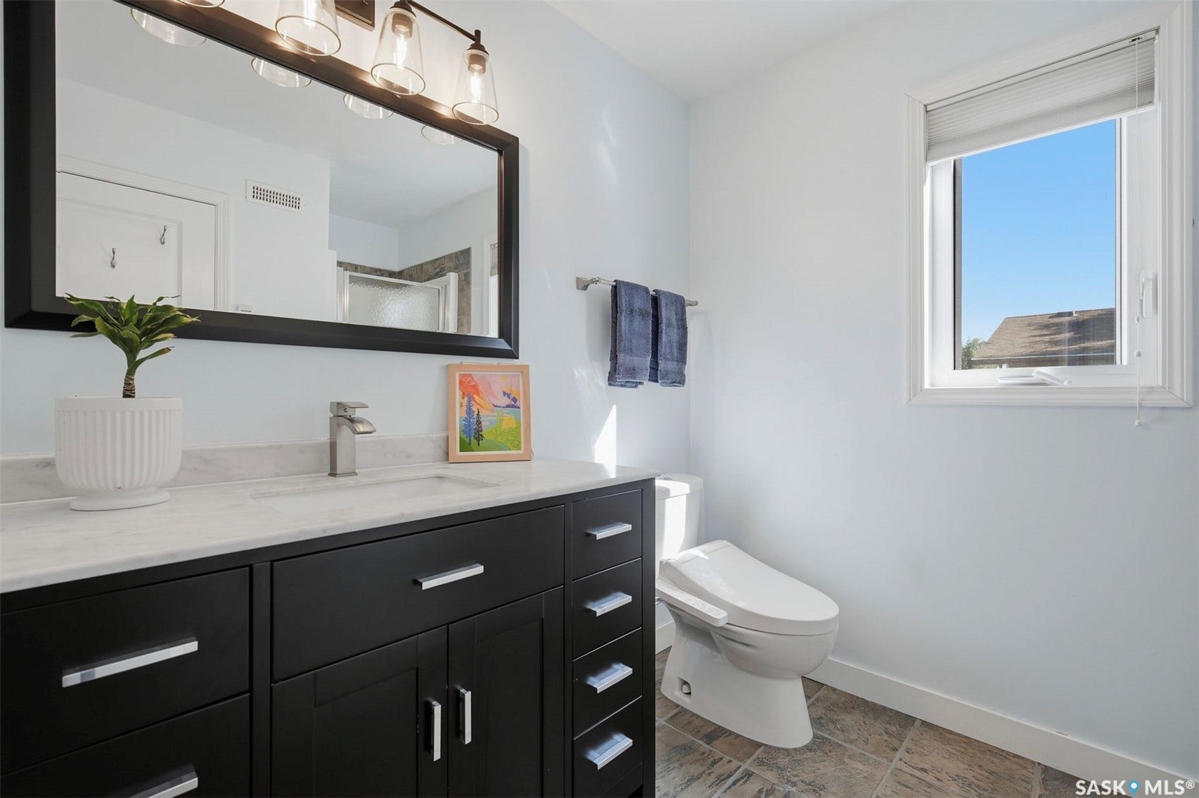 Black vanity with a white countertop and integrated sink, framed mirror with overhead lighting, white walls, and a window with a roller shade