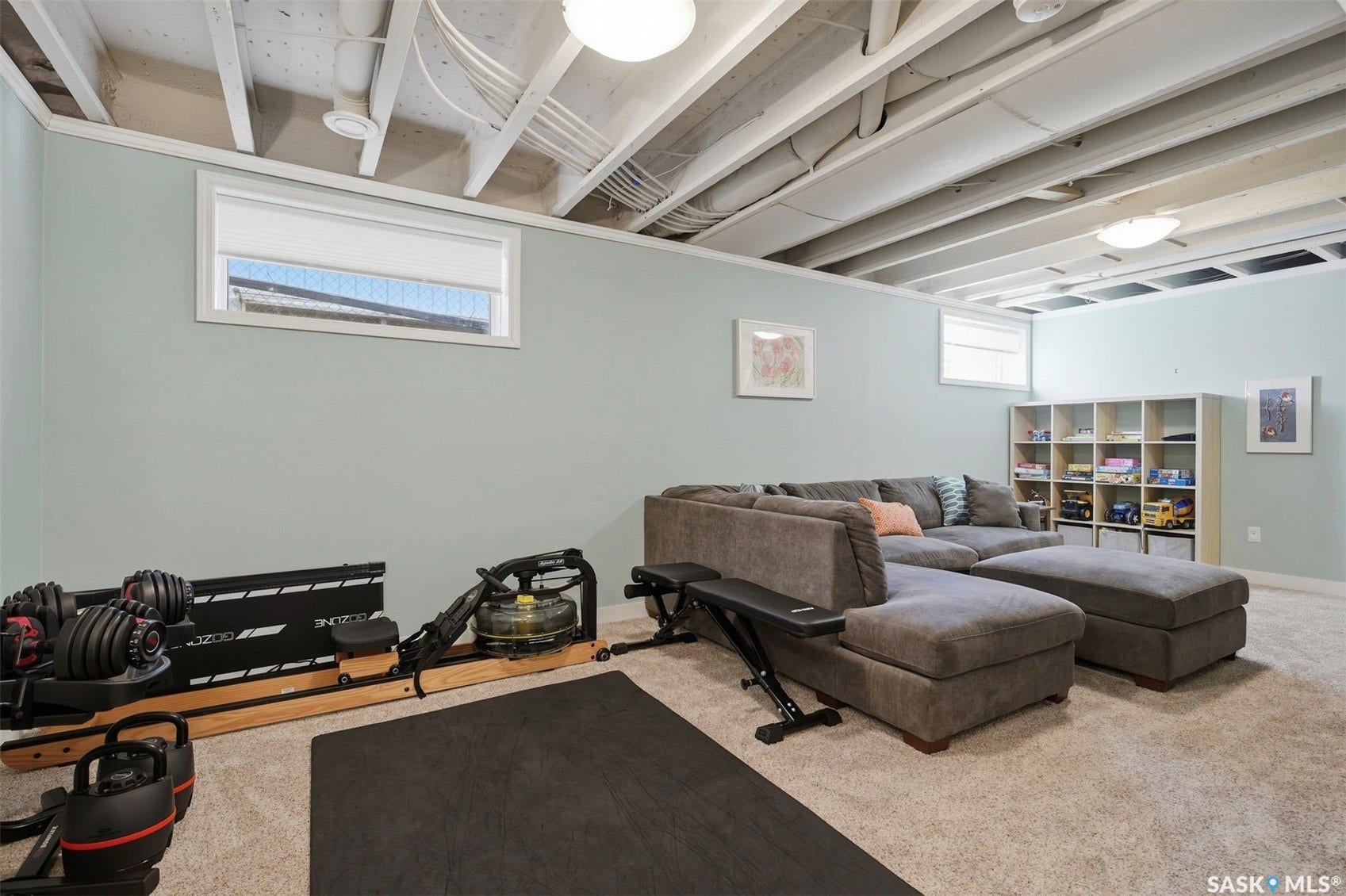 Finished basement space featuring light blue walls, exposed white ceiling joists, and warm neutral carpeting