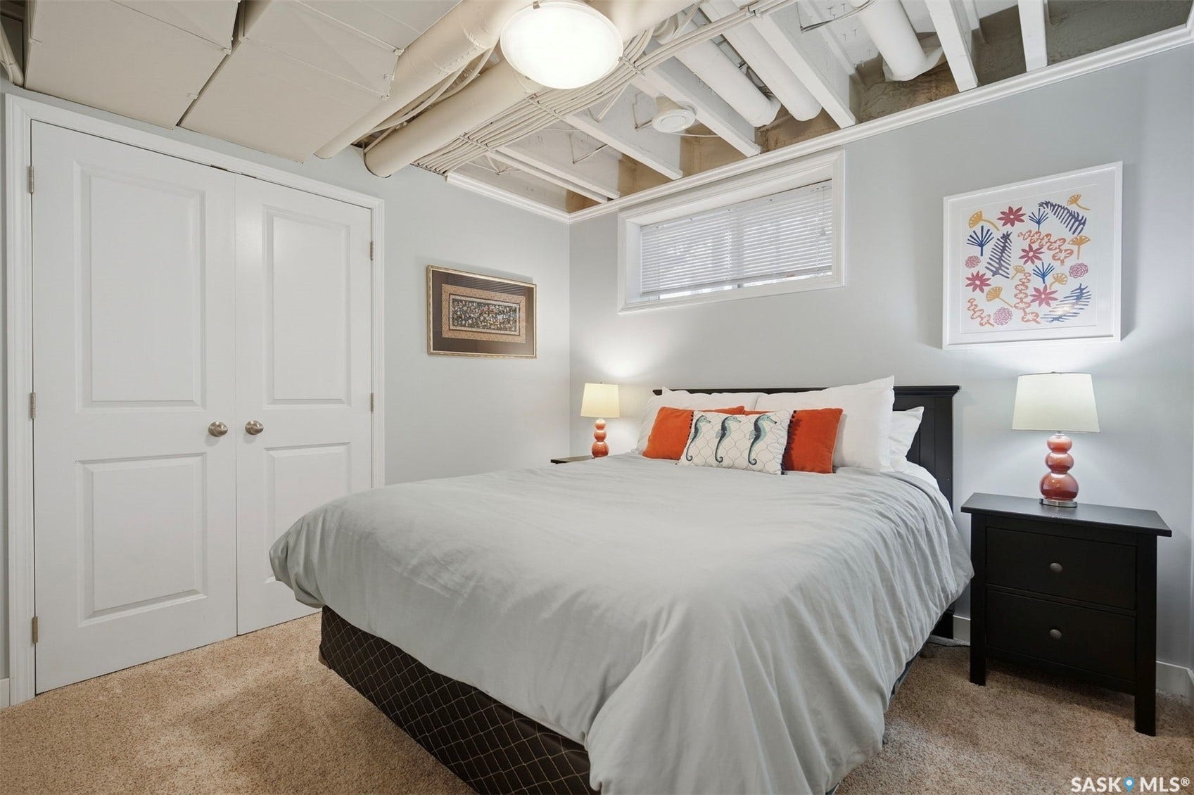 Recessed window with blinds, exposed ceiling beams, built-in ceiling light fixture, white bi-fold closet doors, and light-colored carpet flooring