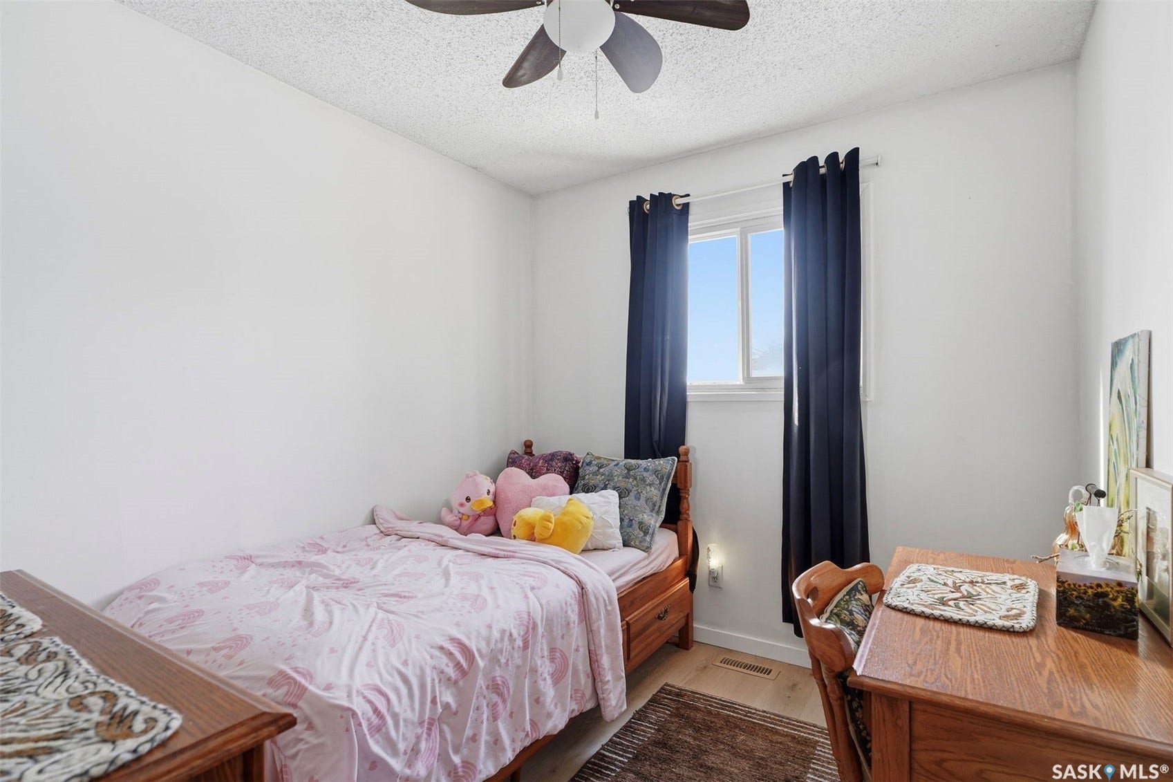 Bright secondary bedroom featuring hardwood flooring, a large window with black grommet curtains providing natural light, ceiling fan with integrated lighting, and a wood-finish desk