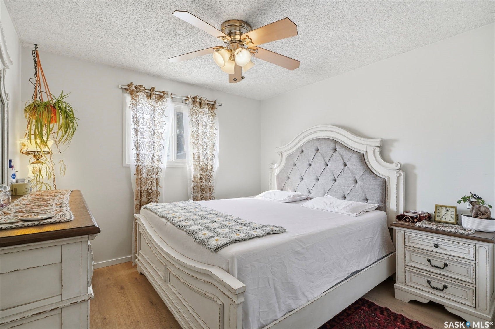 Serene primary bedroom retreat showcasing a white wood bed frame with an upholstered headboard, hardwood flooring, ceiling fan with integrated lighting, sheer curtains framing dual windows, and a coordinating nightstand with drawer storage