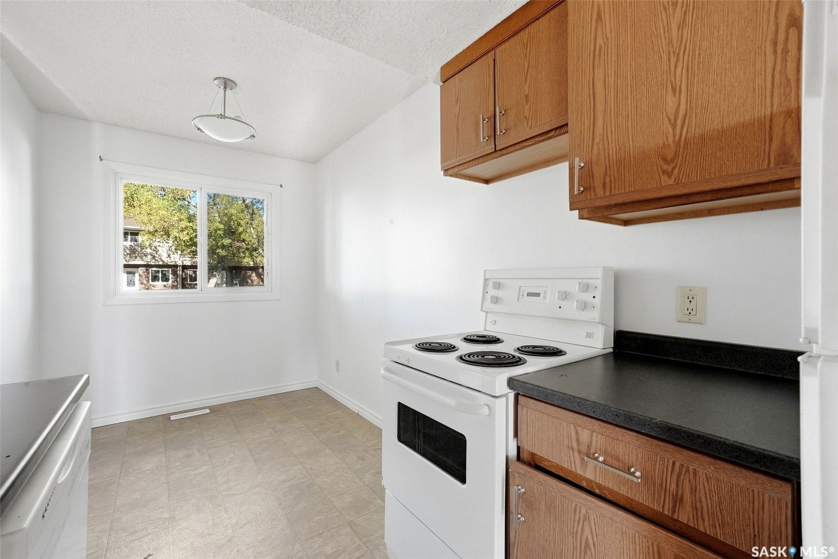 White electric range with a built-in oven, wood-finish cabinetry, dark countertops, a window with a white frame, and a ceiling-mounted light fixture