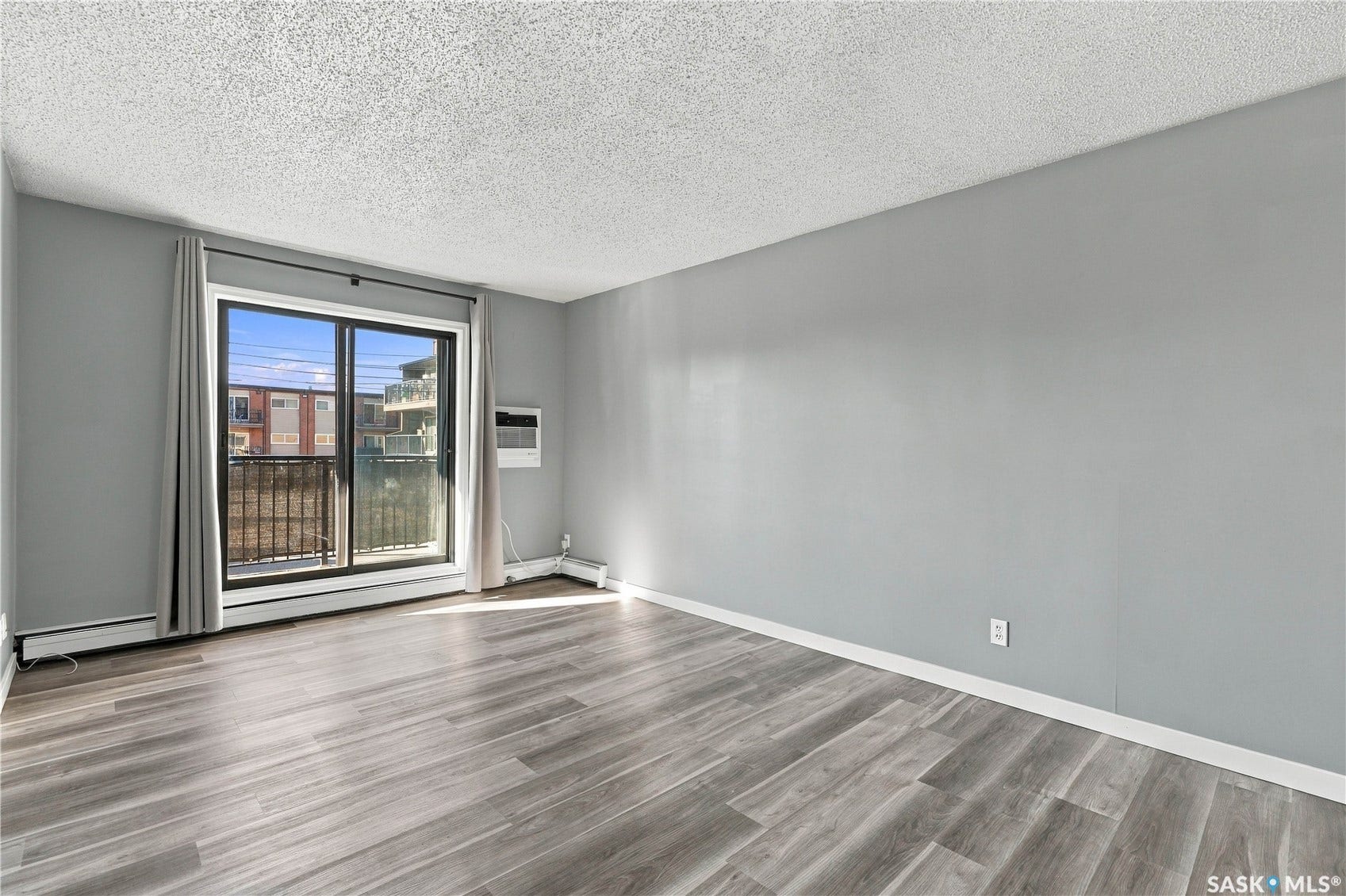 Spacious interior with wood-finish flooring, light gray wall paint, and a sliding glass door leading to a balcony with a black metal railing