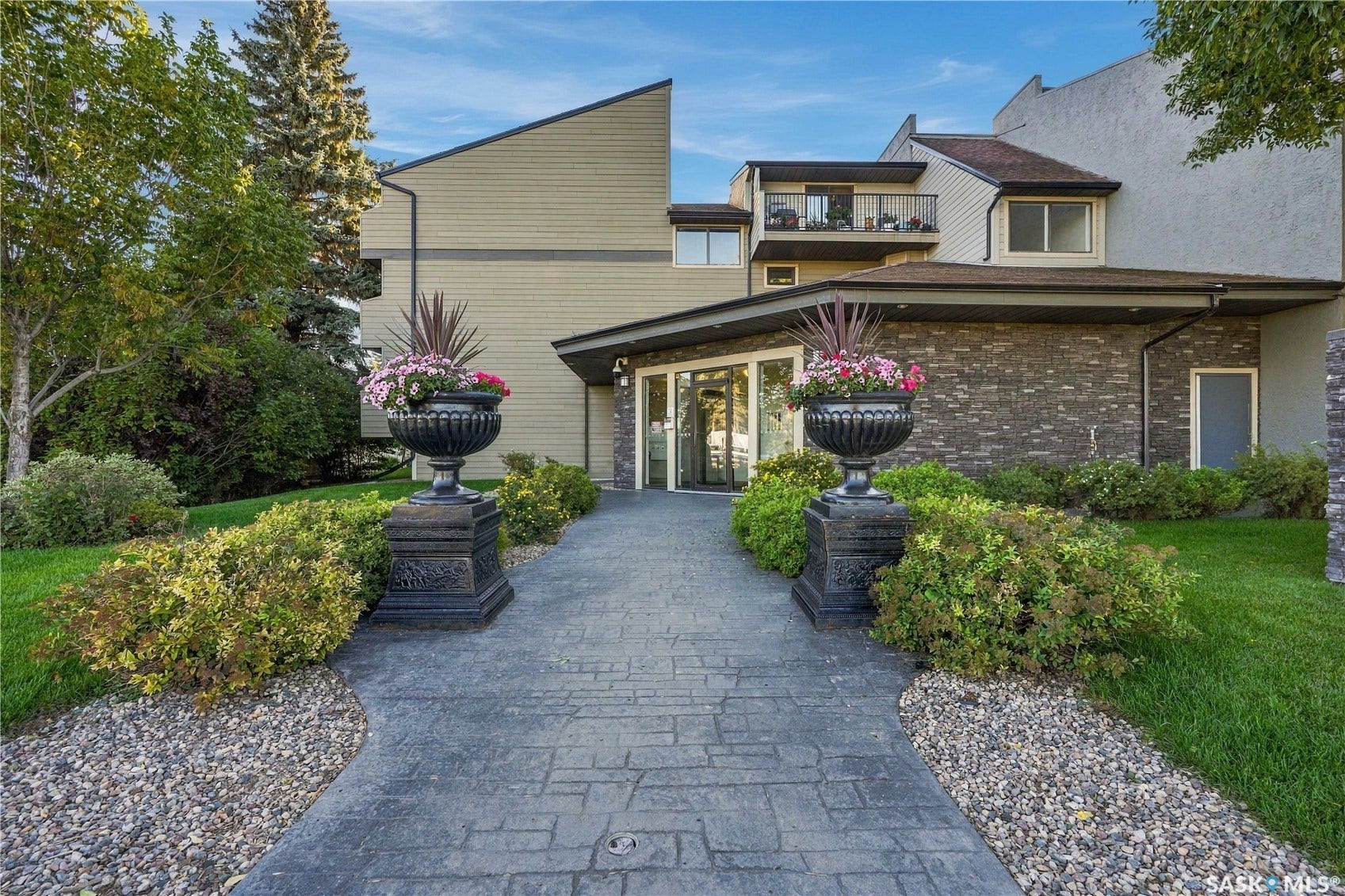 Patterned paver walkway leading to a glass-paneled entry, framed by mature landscaping and decorative planters