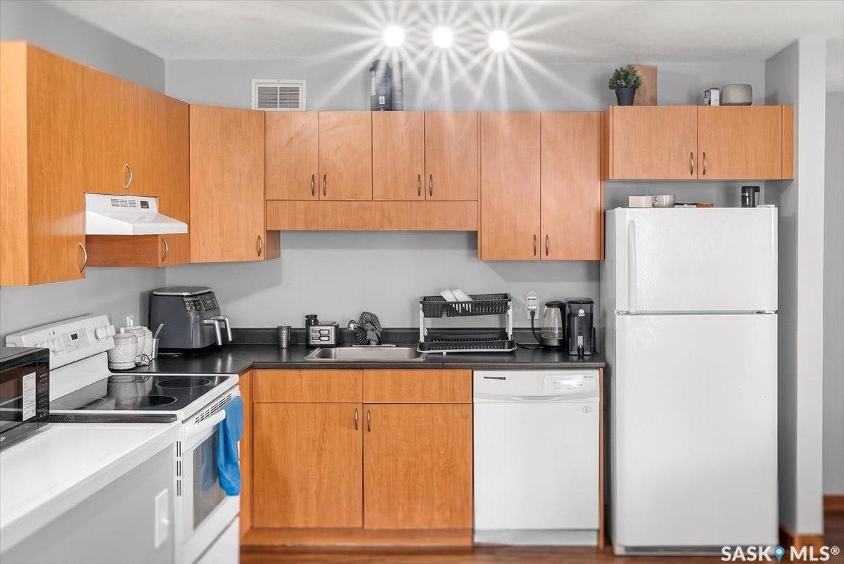 5051 James Hill Road, #107, Regina SK S4W 0B9 Kitchen featuring wood-finish cabinetry, dark countertops, a built-in range hood, and a white dishwasher
