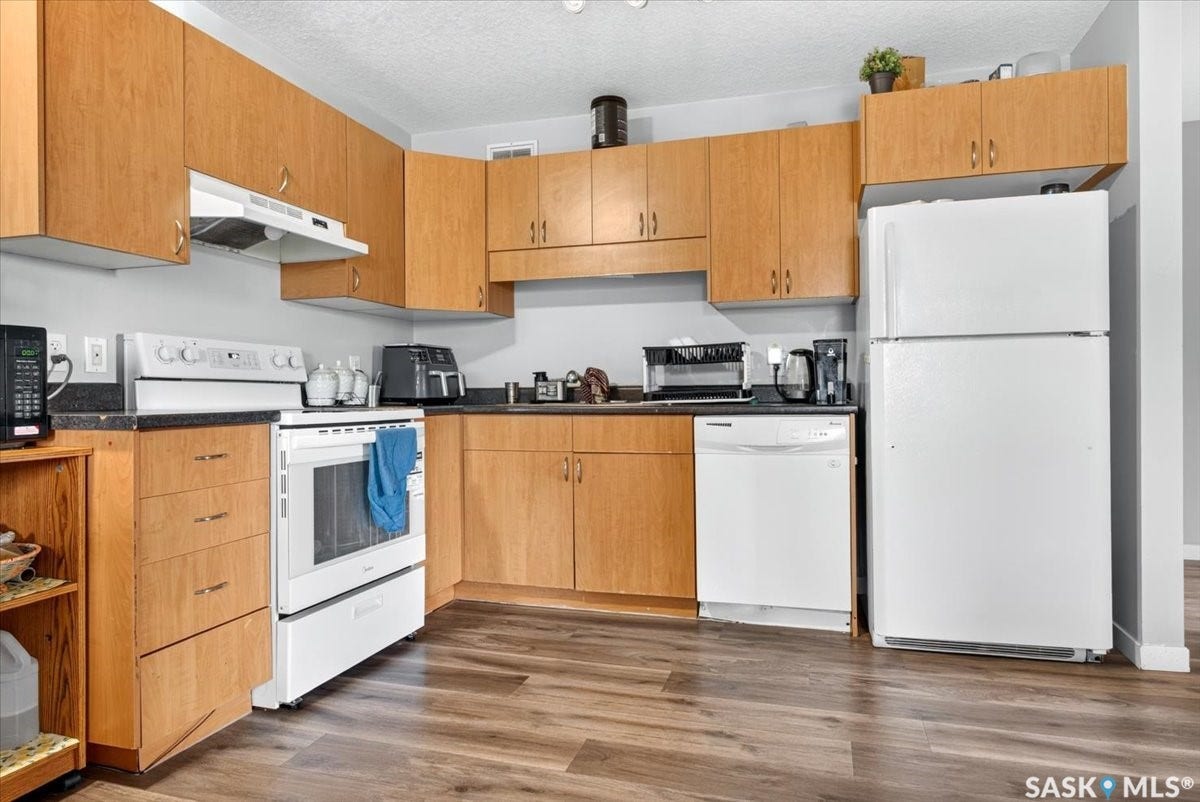 5051 James Hill Road, #107, Regina SK S4W 0B9 Kitchen featuring wood-finish cabinetry, dark countertops, a white electric range, a white dishwasher, and a white refrigerator