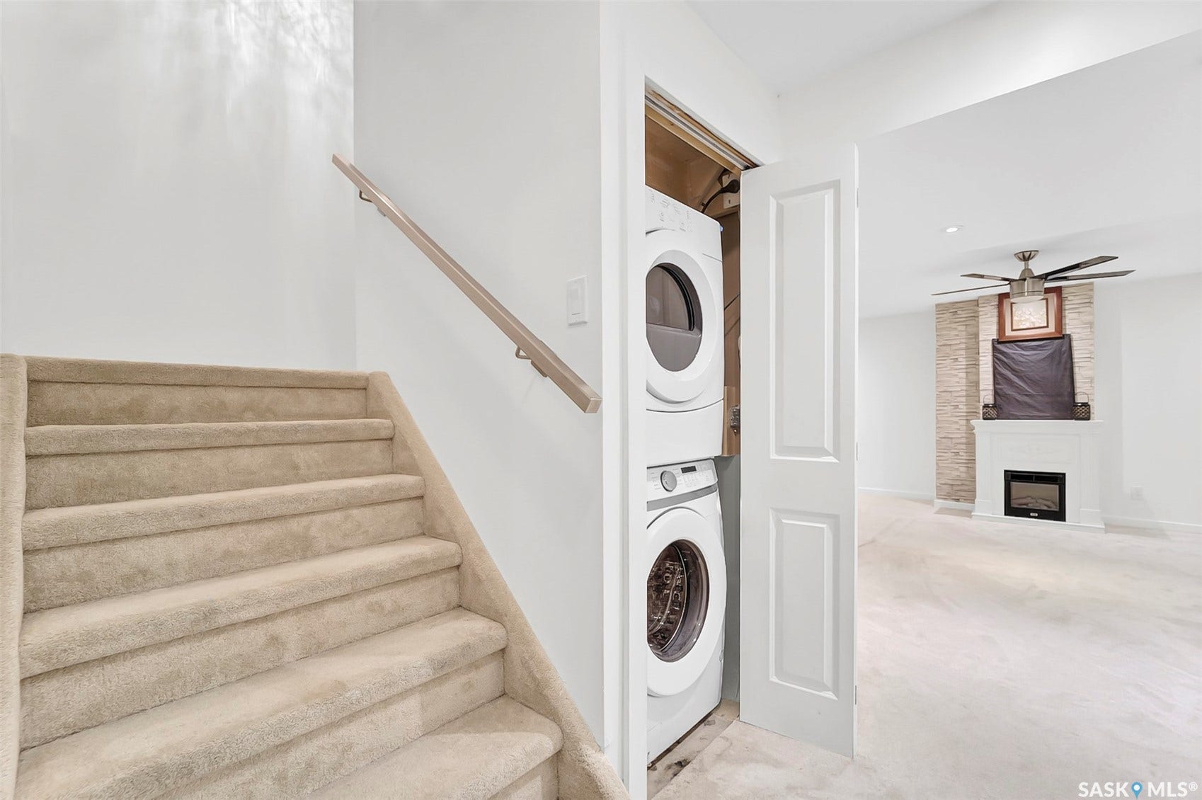 Carpeted staircase with wooden handrail, stacked washer and dryer, fireplace with stone surround, and ceiling fan