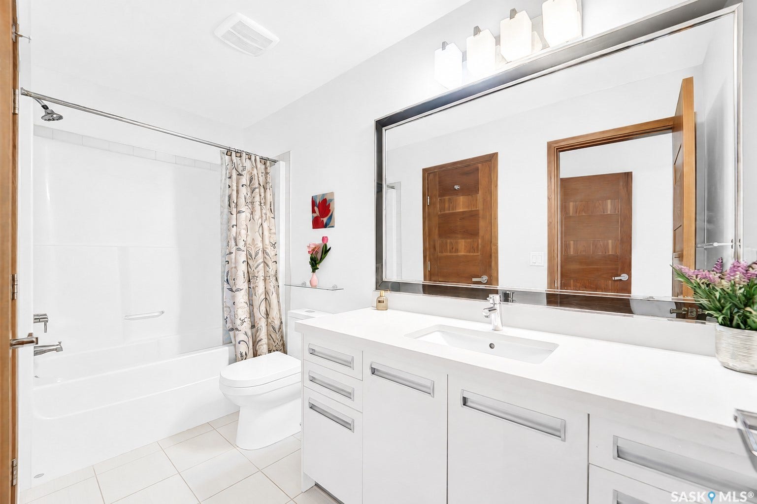 White vanity with a rectangular undermount sink, a large mirror with integrated lighting, a white bathtub with a shower, and white tile flooring