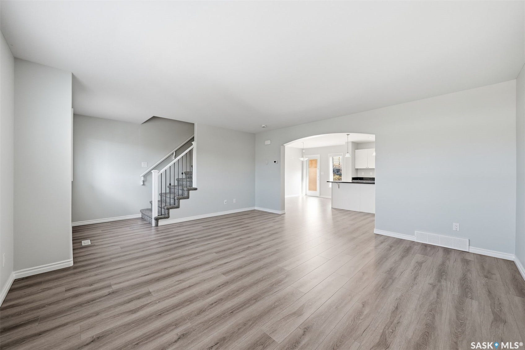 Spacious interior with wood-finish flooring, a built-in staircase with white railings, and an arched doorway leading to a kitchen with white cabinetry