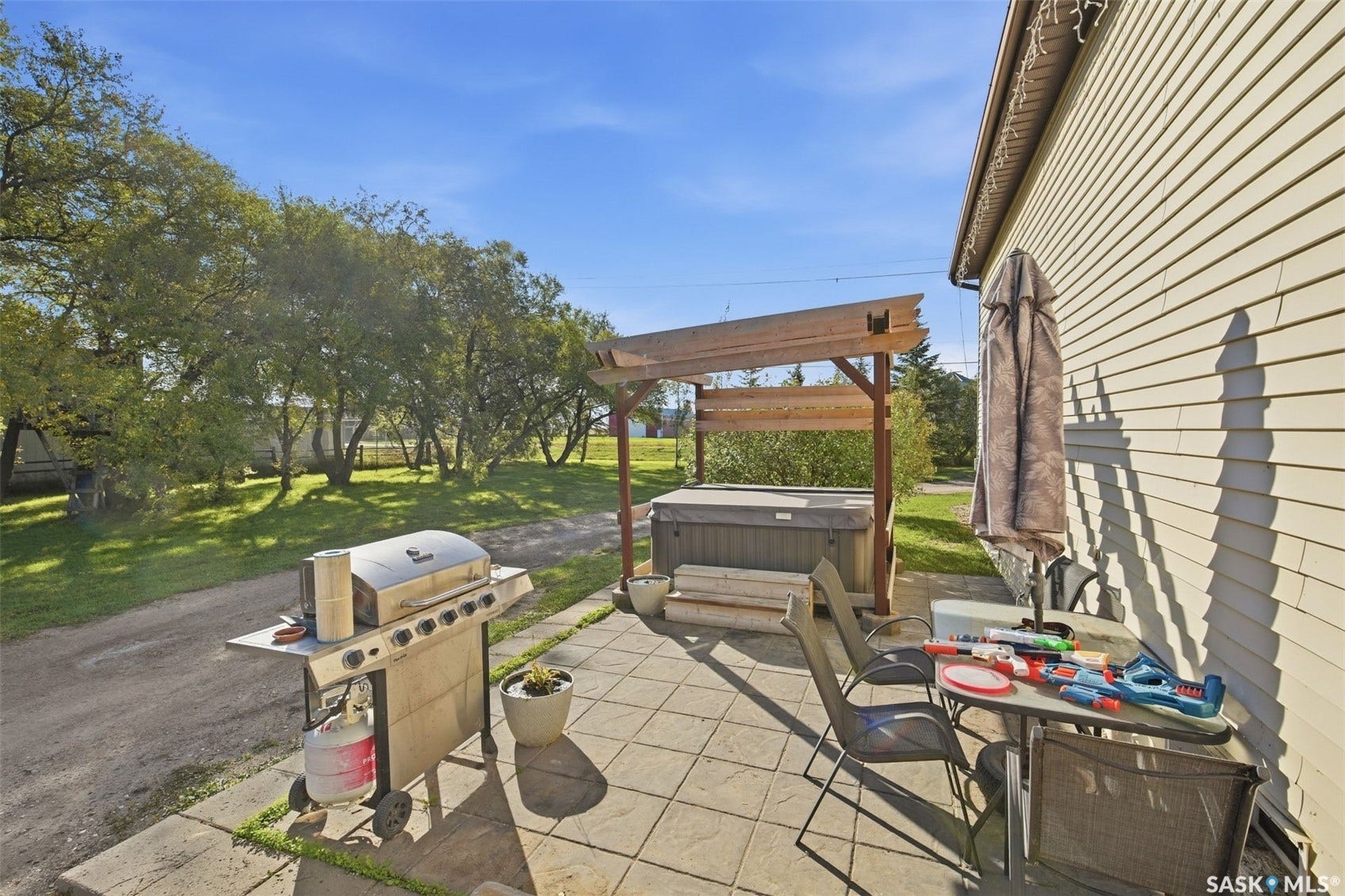 Paved patio area featuring a hot tub with an overhead pergola, and a grassy expanse with mature trees