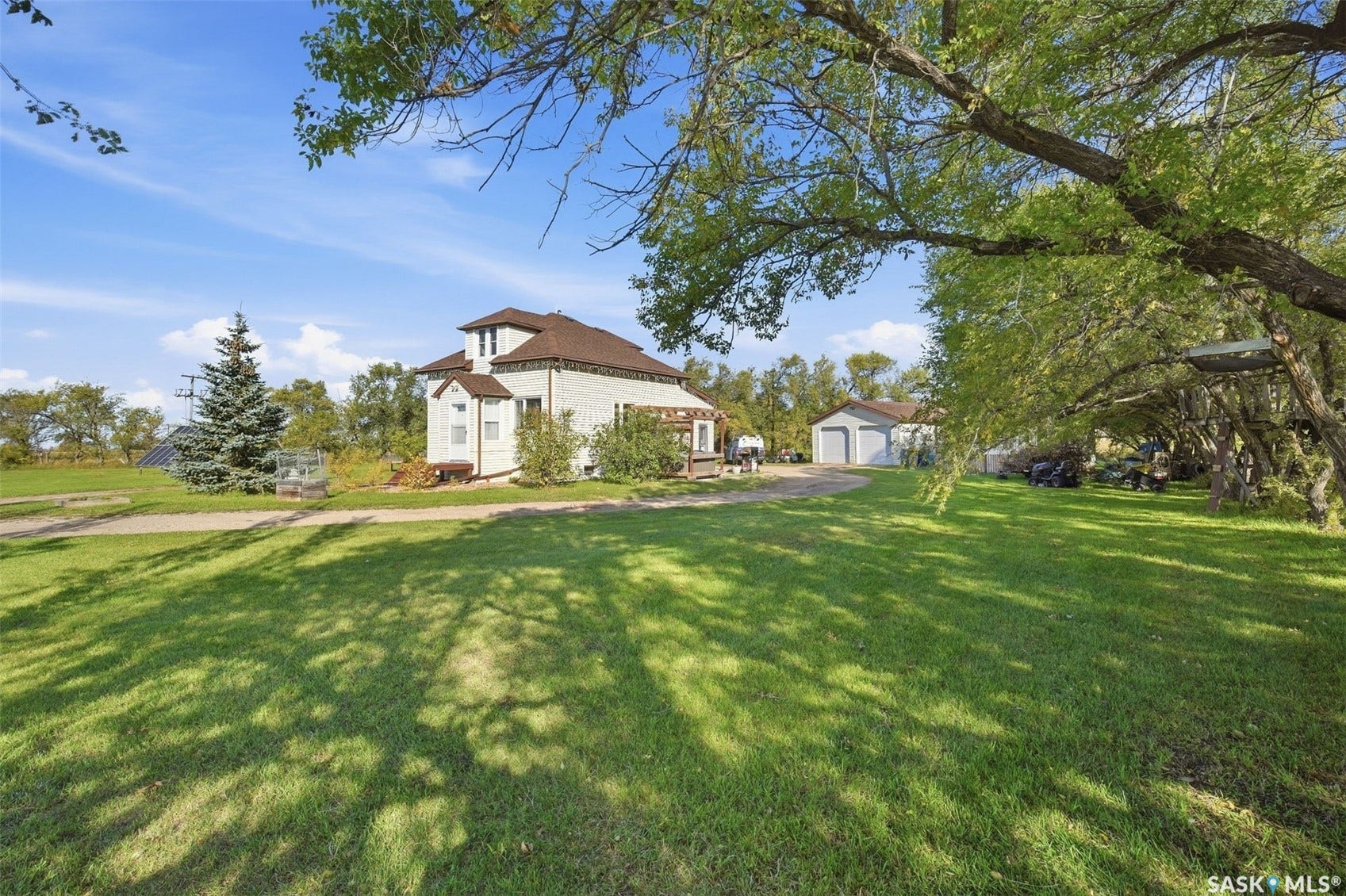 Expansive green lawn, mature trees, white exterior home with brown roof, detached garage with two bay doors, and a paved driveway