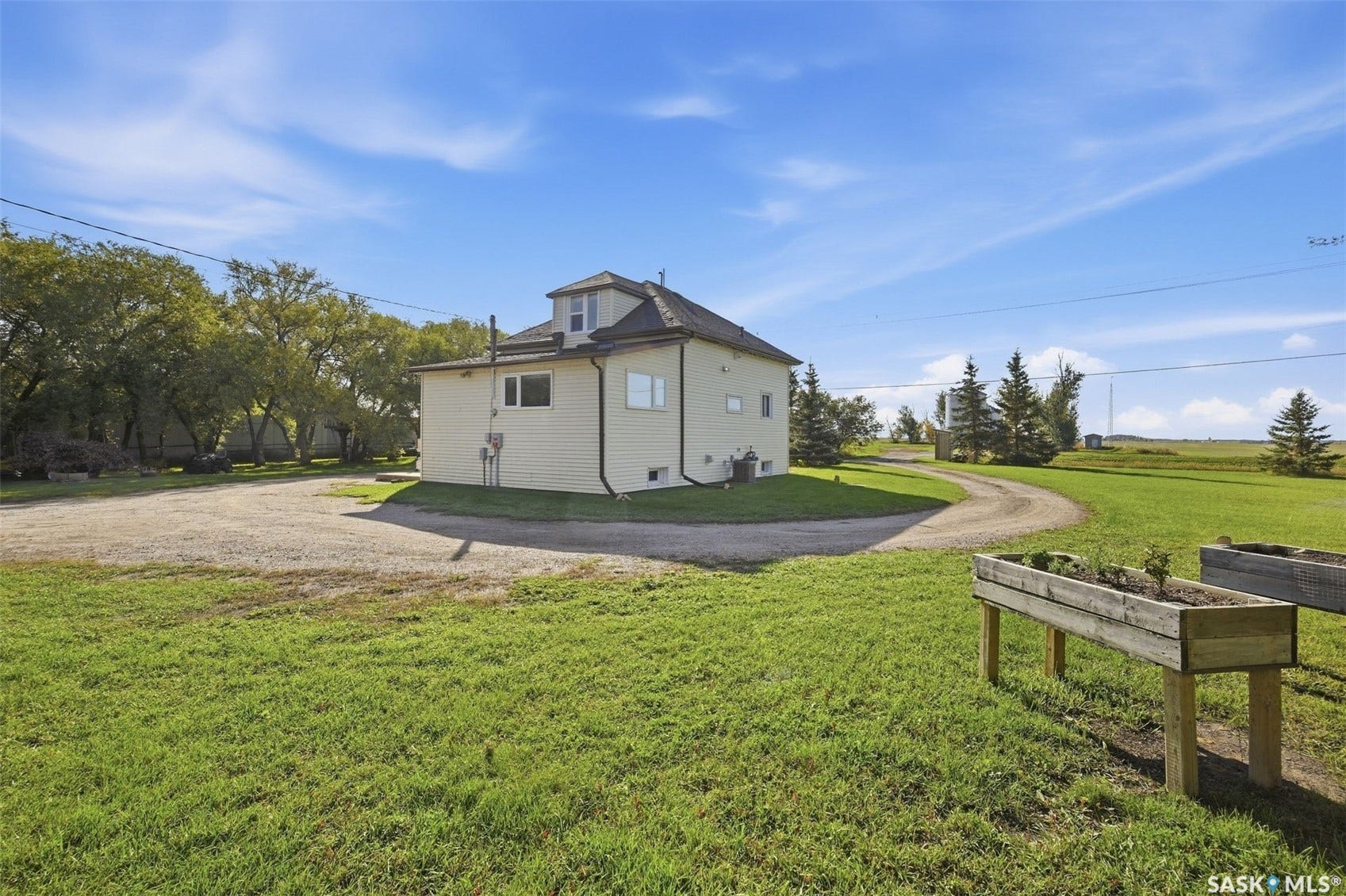 Expansive grassy yard with mature trees, gravel driveway, and raised garden beds