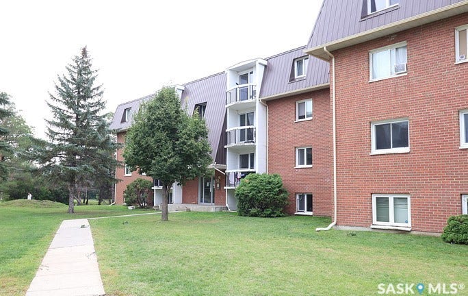 Brick exterior, metal mansard roof, balconies with white railings, mature trees, and a concrete walkway