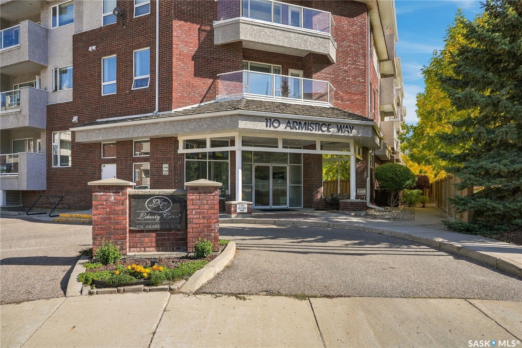 Brick and stucco exterior, prominent building signage, glass balcony railings, mature evergreen trees, and a decorative brick sign