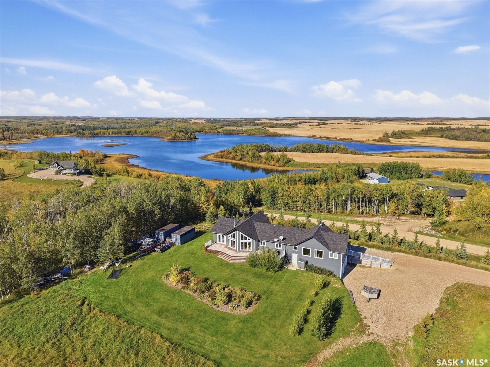 Expansive property featuring a grey siding exterior, a dark shingle roof, a large unpaved parking area, and a significant body of water in the background