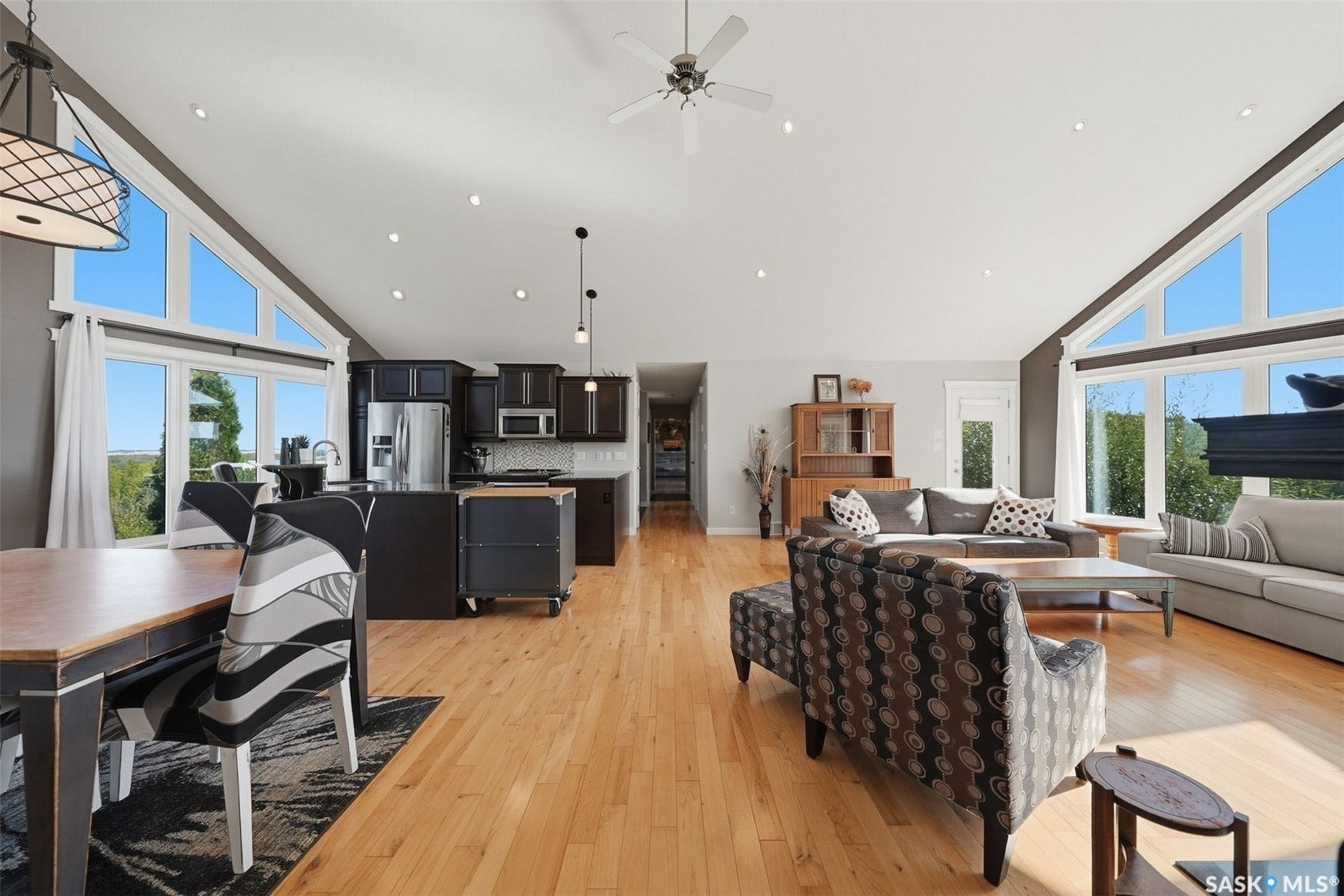 Vaulted ceiling with recessed lighting, light wood-finish flooring, full-height windows, a ceiling fan, and a kitchen with dark cabinetry and stainless steel appliances