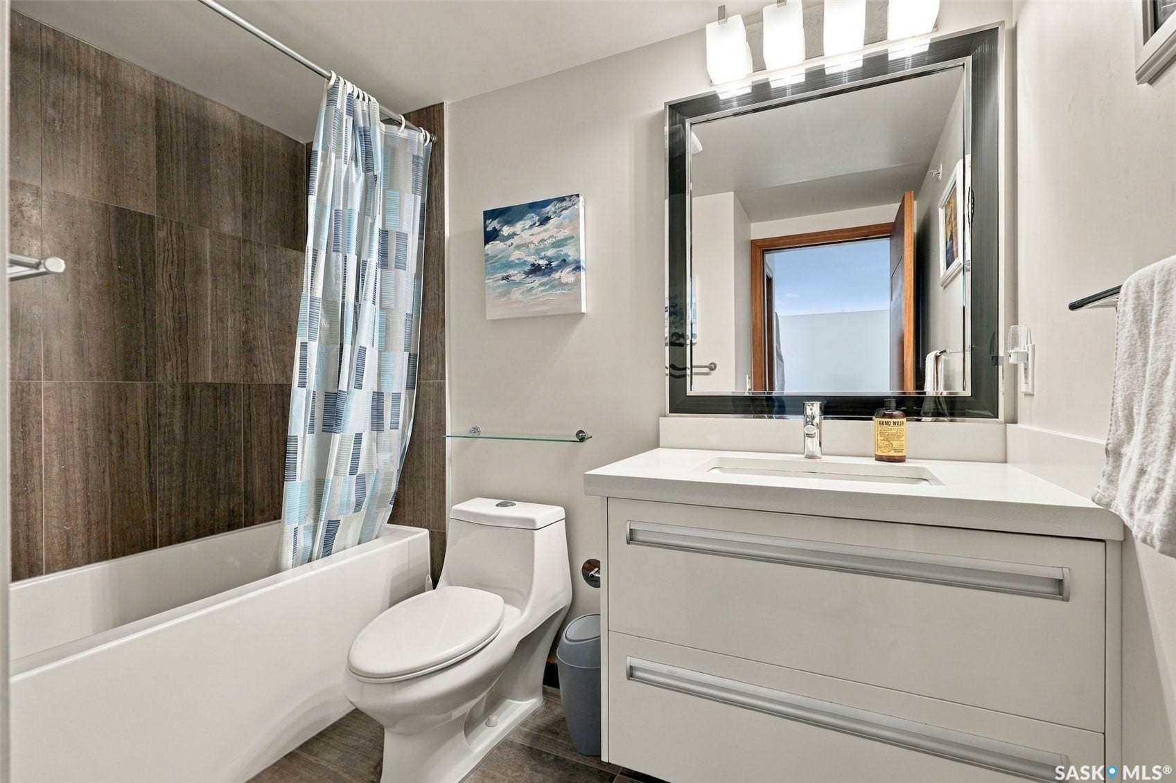 White vanity with integrated sink and chrome faucet, framed mirror with vanity lighting, full bathtub with tiled surround, and a toilet with an enclosed tank