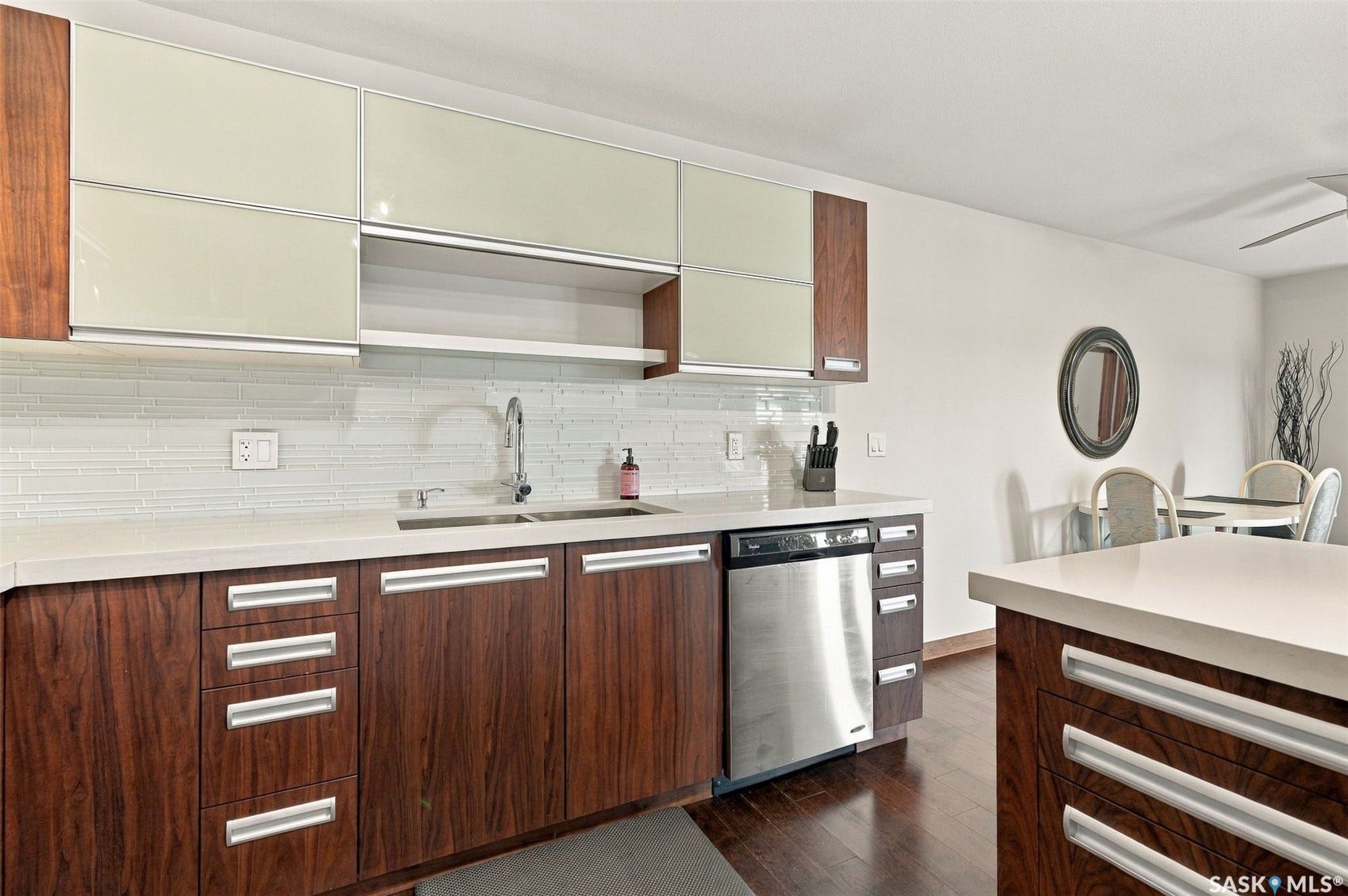 Wood-finish lower cabinetry, light-toned upper cabinetry, stainless steel sink, white countertop, and tiled backsplash