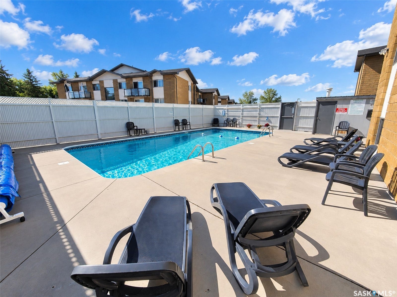 Concrete pool deck surrounding a rectangular swimming pool, featuring a white privacy fence and a brick-faced building in the background
