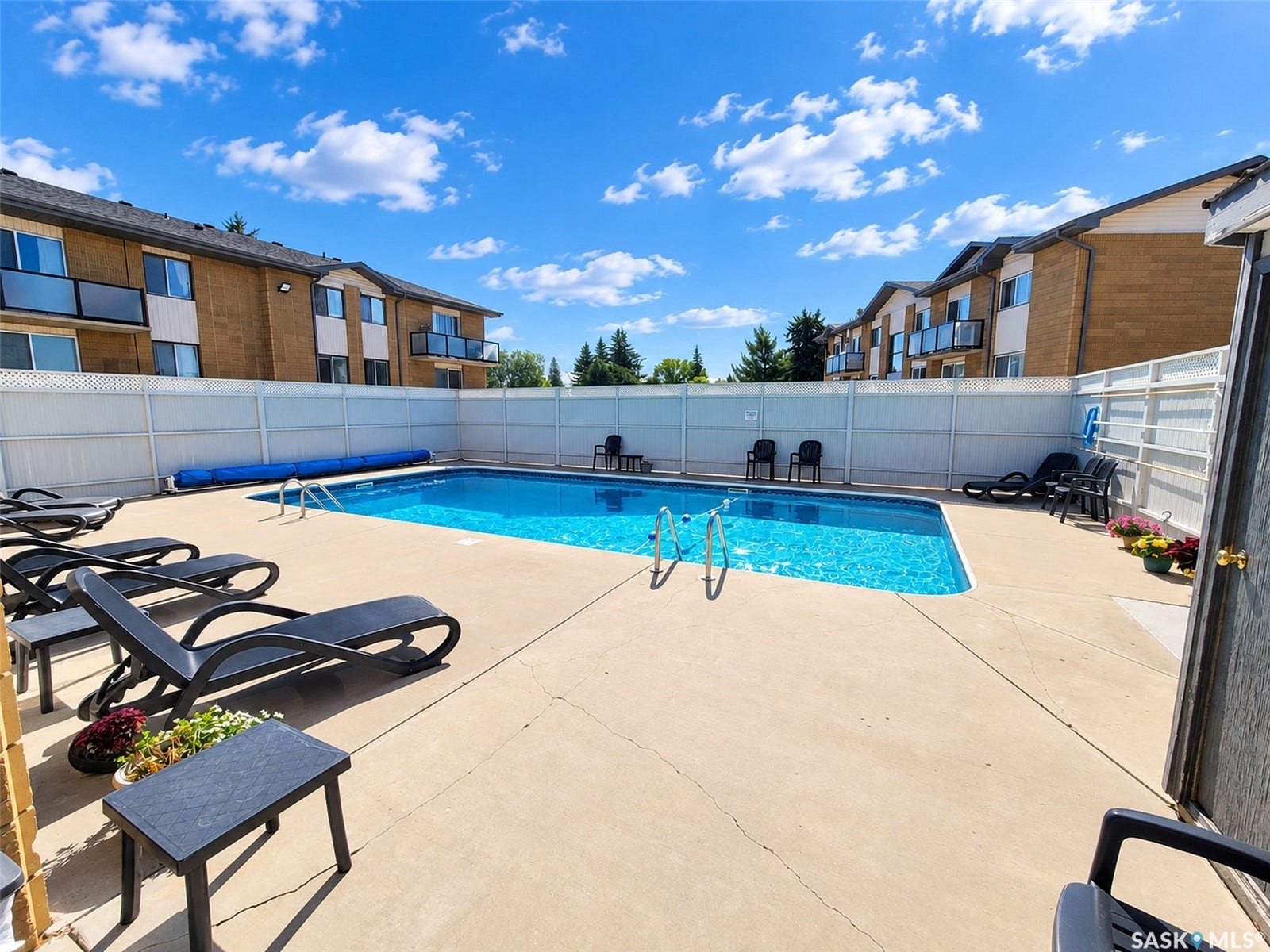 Outdoor swimming pool with concrete decking, surrounded by white privacy fencing, with multi-story buildings in the background