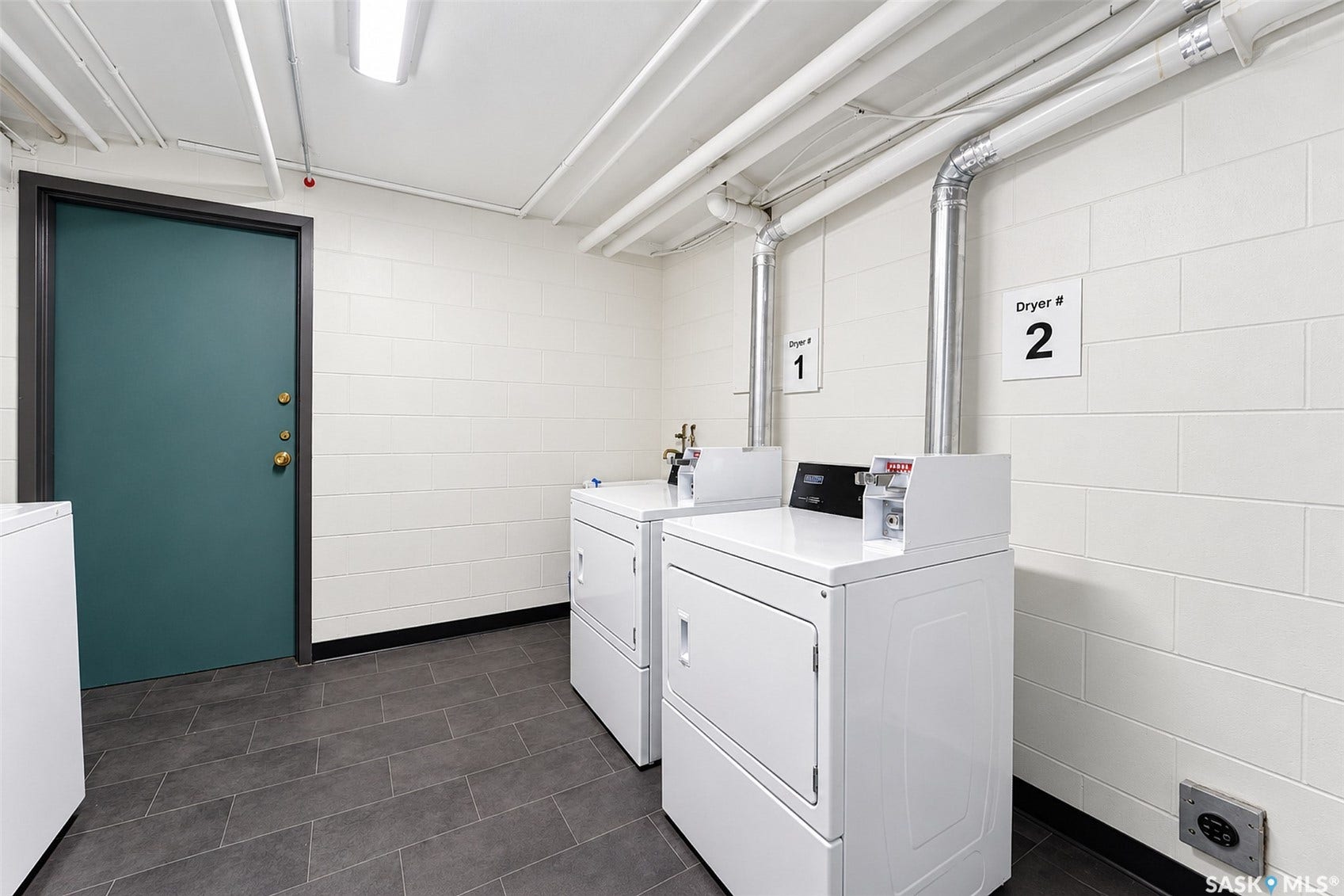 Laundry facility with two white dryers, exposed ceiling pipes, and grey tile flooring
