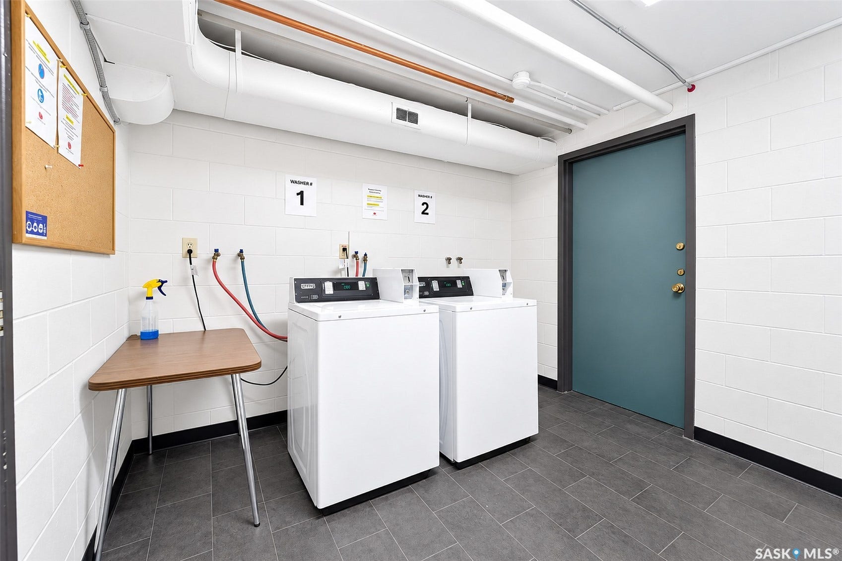 Laundry area with two washing machines, white block walls, dark gray floor tiling, and exposed ceiling utilities
