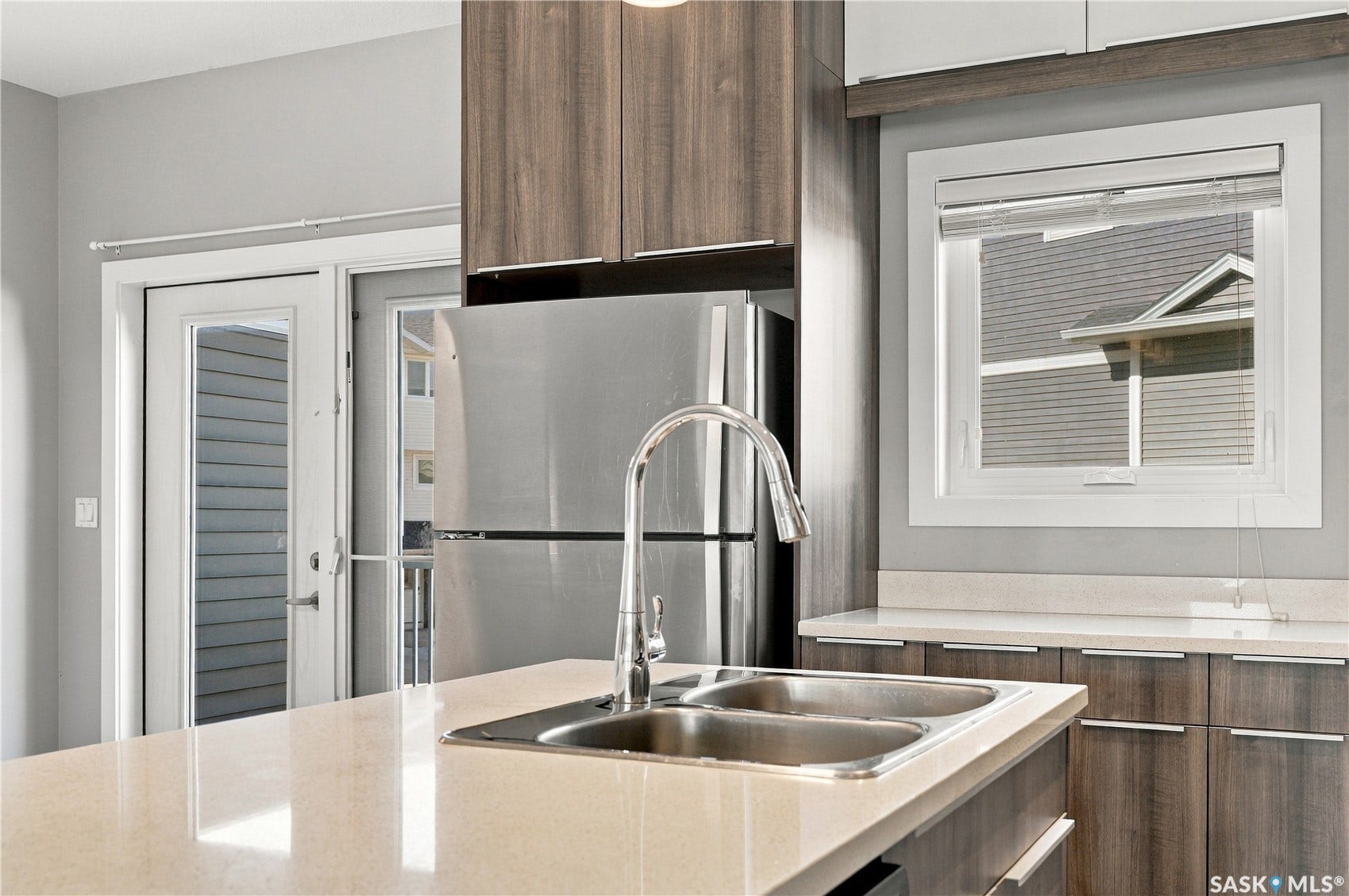 Stainless steel refrigerator, dual basin sink with gooseneck faucet, light-toned countertops, and wood-finish cabinetry