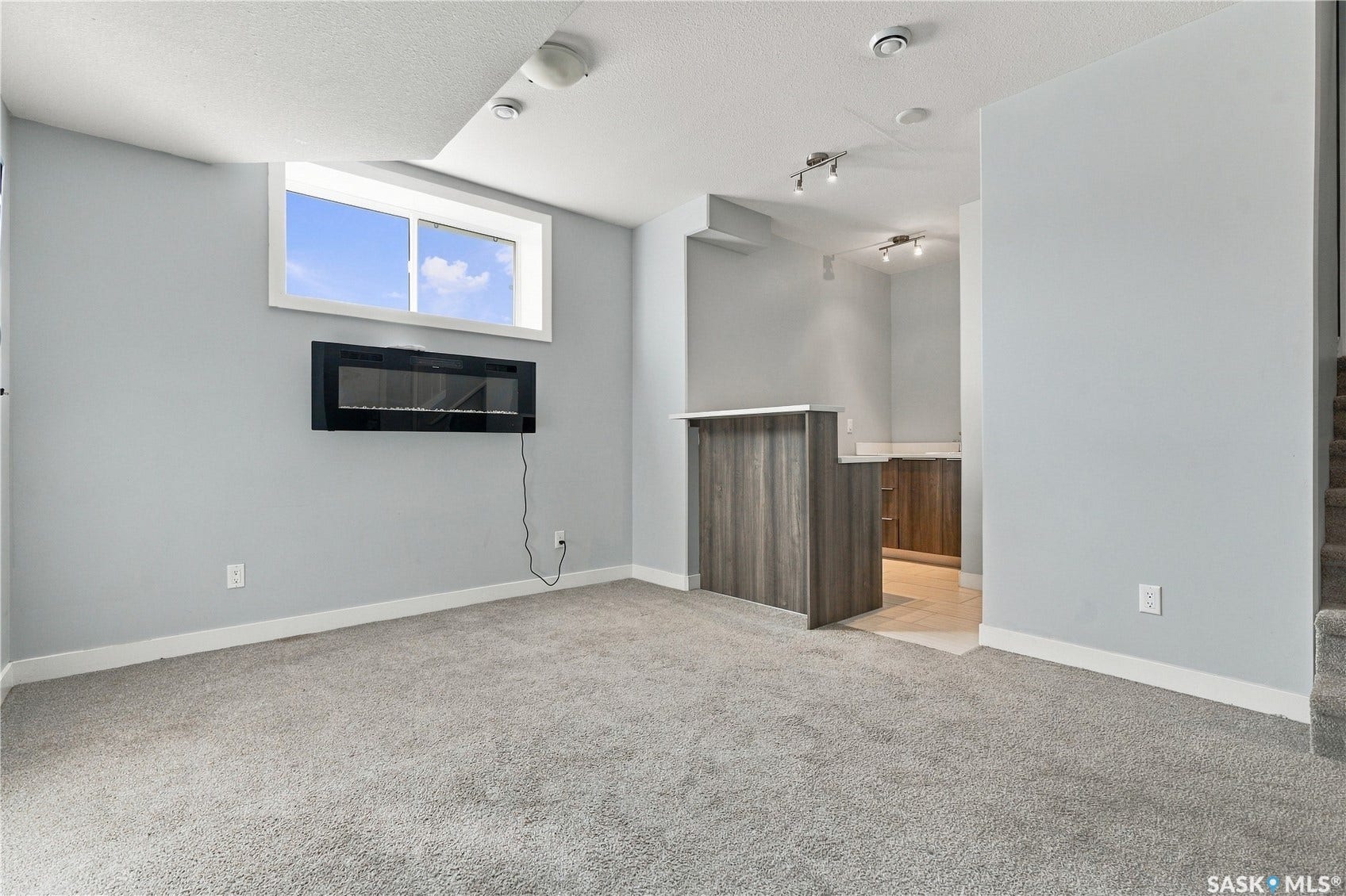 Recessed window, wall-mounted electric fireplace, carpeted flooring, wood-finish bar with a light-toned countertop, and track lighting