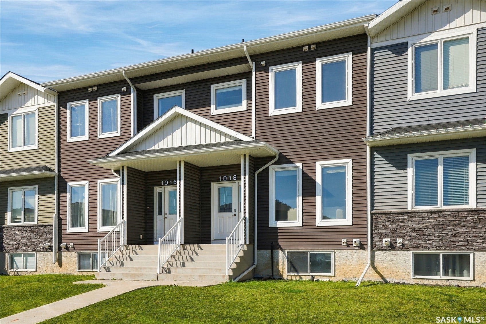 Contemporary townhome featuring a clean-lined facade with mixed siding and stone accents, expansive multi-pane windows, a covered front entry with white railings, and a manicured lawn with mature landscaping